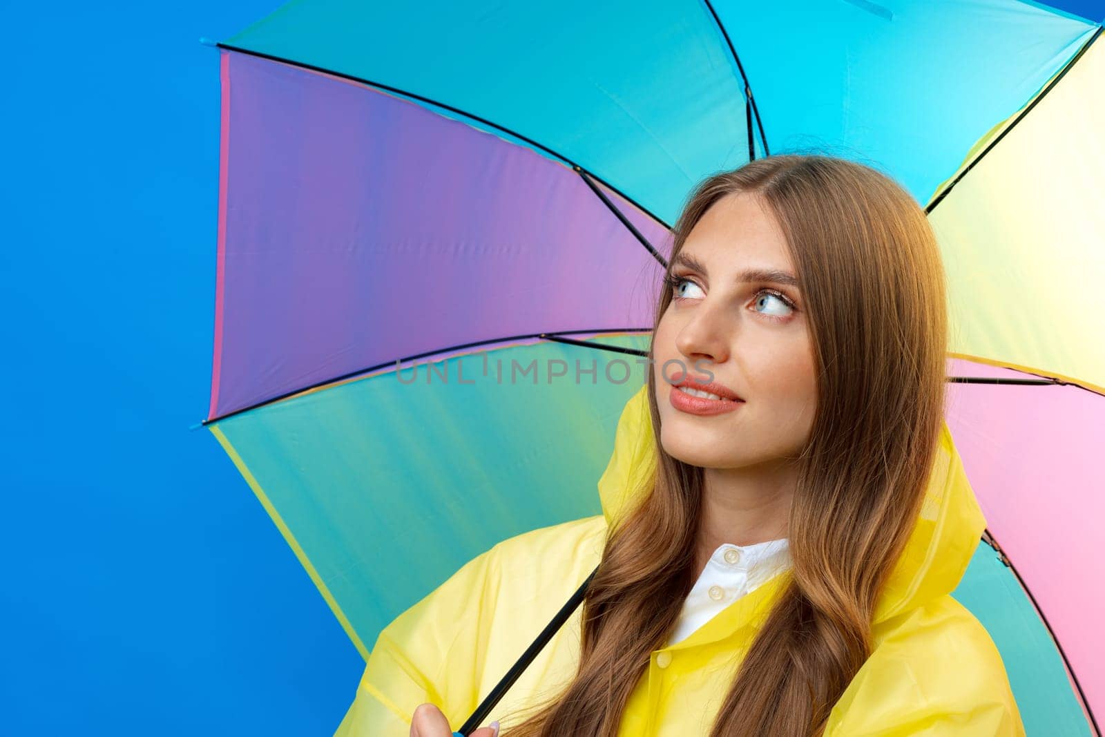 Young woman in yellow raincoat with rainbow umbrella against blue background in studio, close up