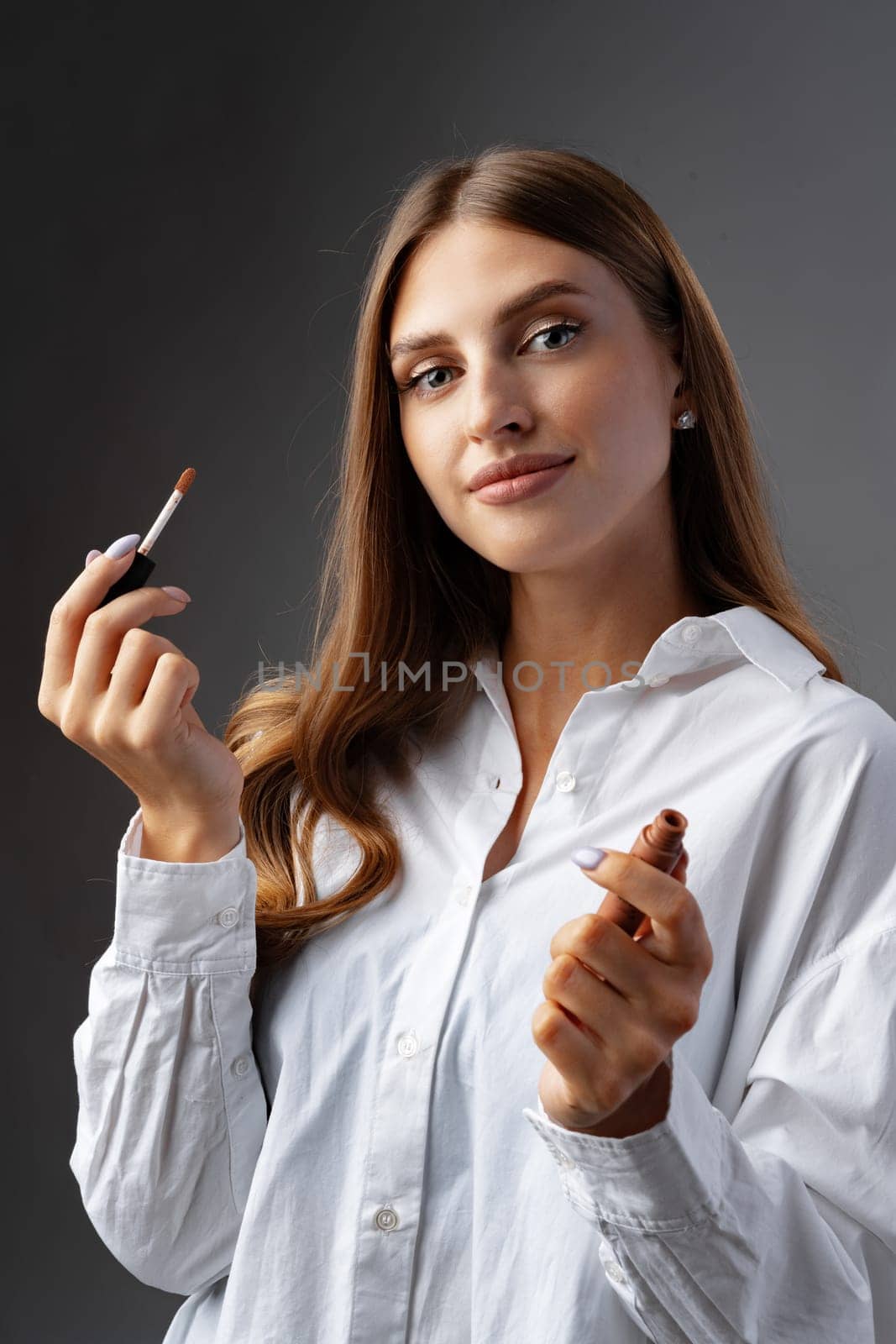 Cute young woman applying lip gloss against gray background in studio