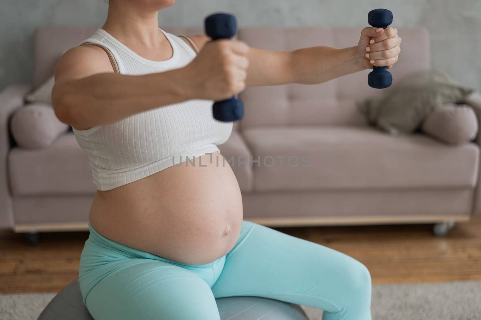 Pregnant woman doing exercises with dumbbells while sitting on a fitness ball at home. Close-up of a pregnant belly