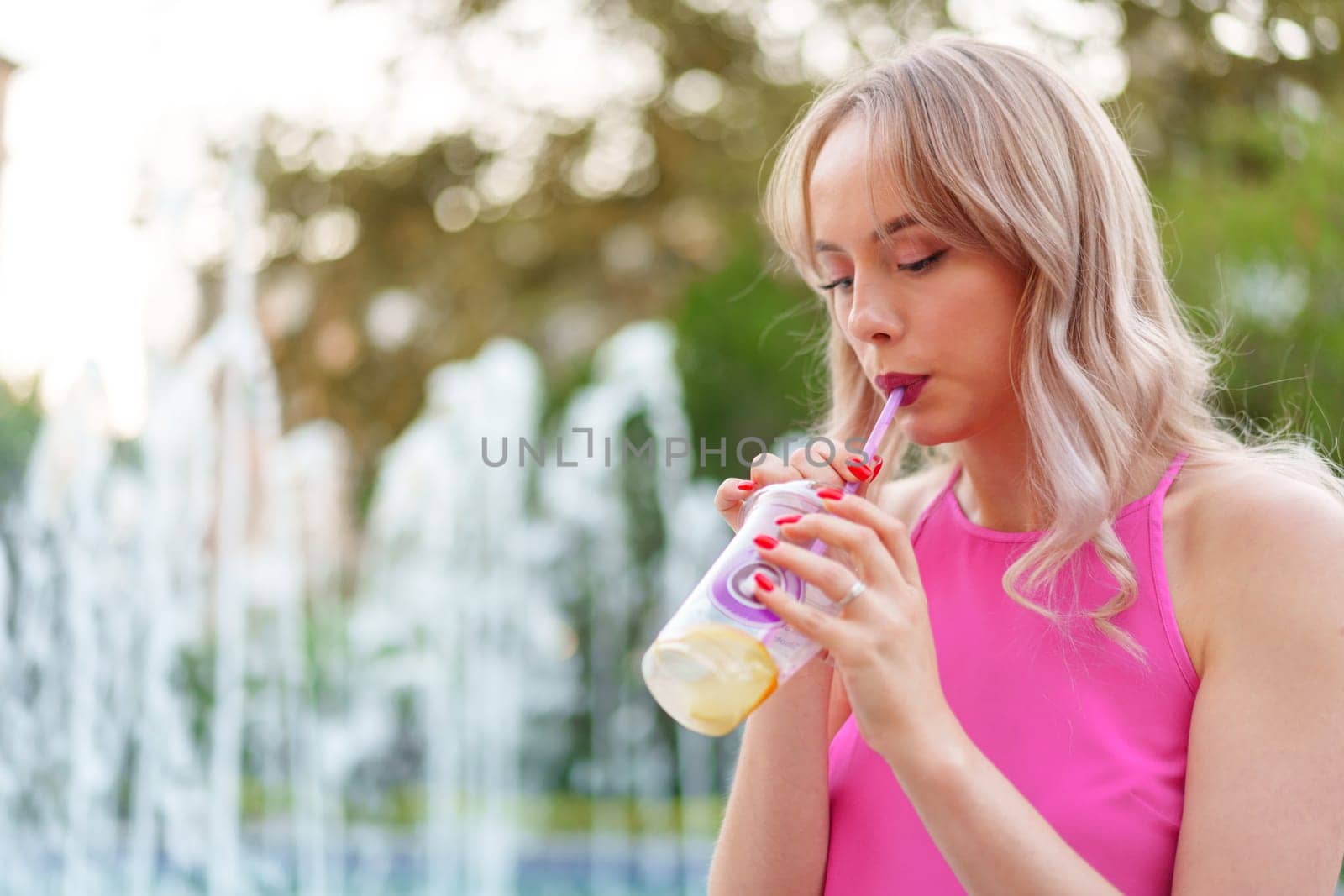 Close up of blonde young woman drinking juice in the street near fountain