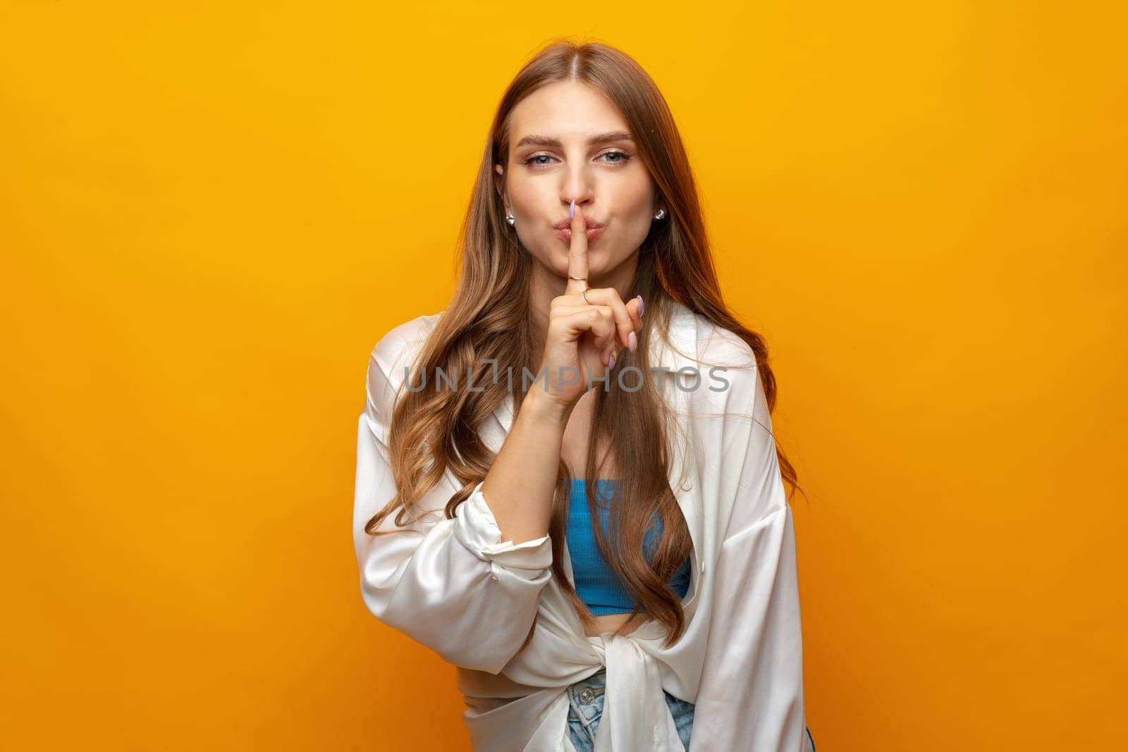 Pretty young woman holding finger on lips and showing silence sign on yellow background close up