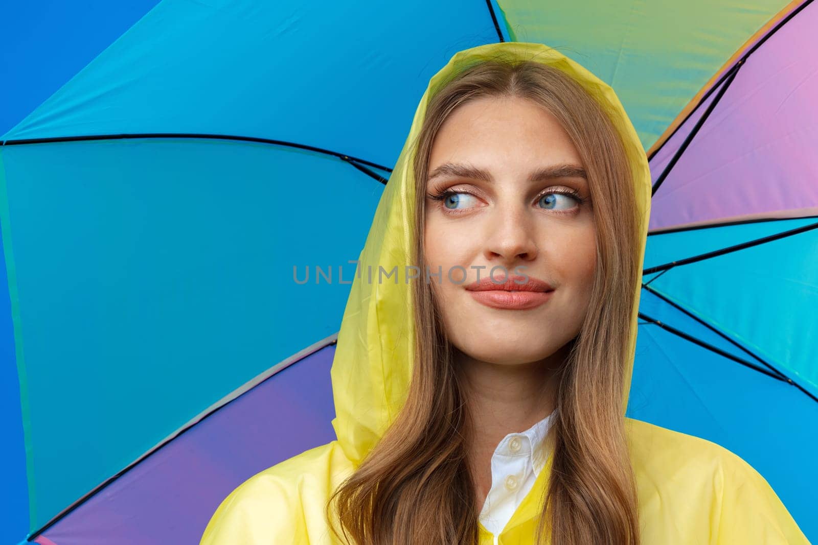 Young woman in yellow raincoat with rainbow umbrella against blue background in studio, close up