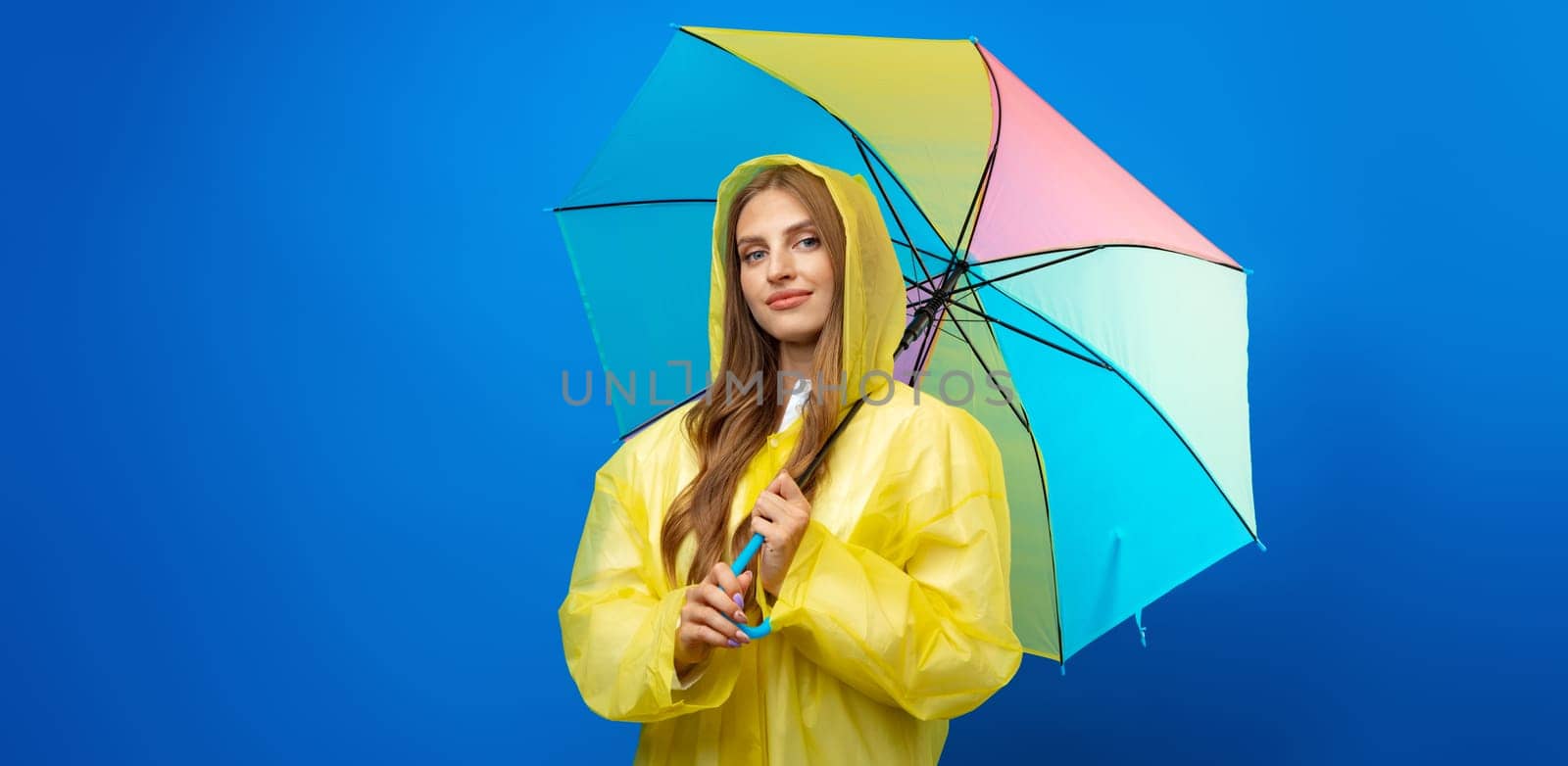 Young woman in yellow raincoat with rainbow umbrella against blue background in studio, close up