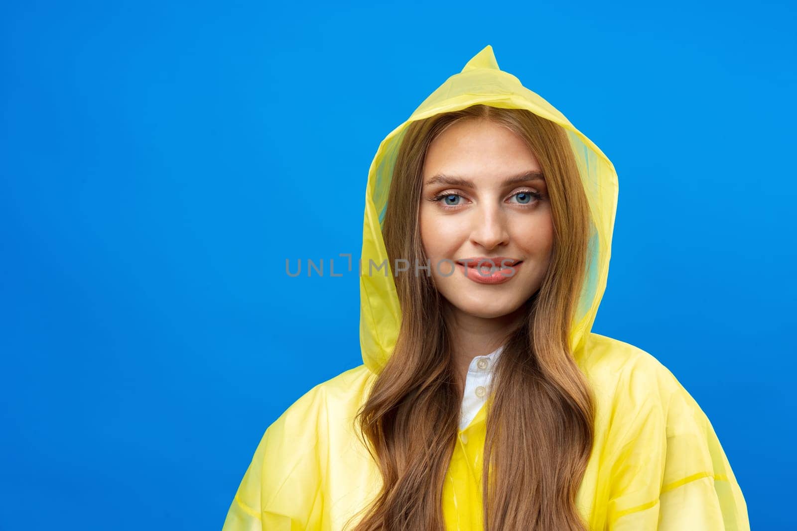Young blonde woman wearing yellow raincoat smiling at camera over blue background in studio, close up
