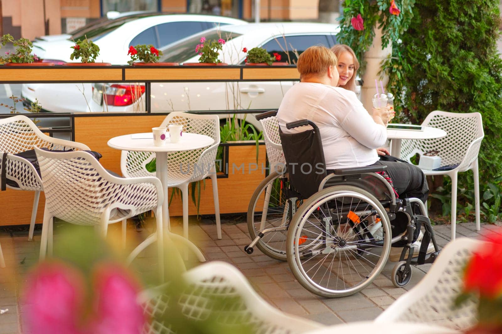 Young daughter and her mother in wheelchair sitting at the table in cafe with drinks and having fun, close up