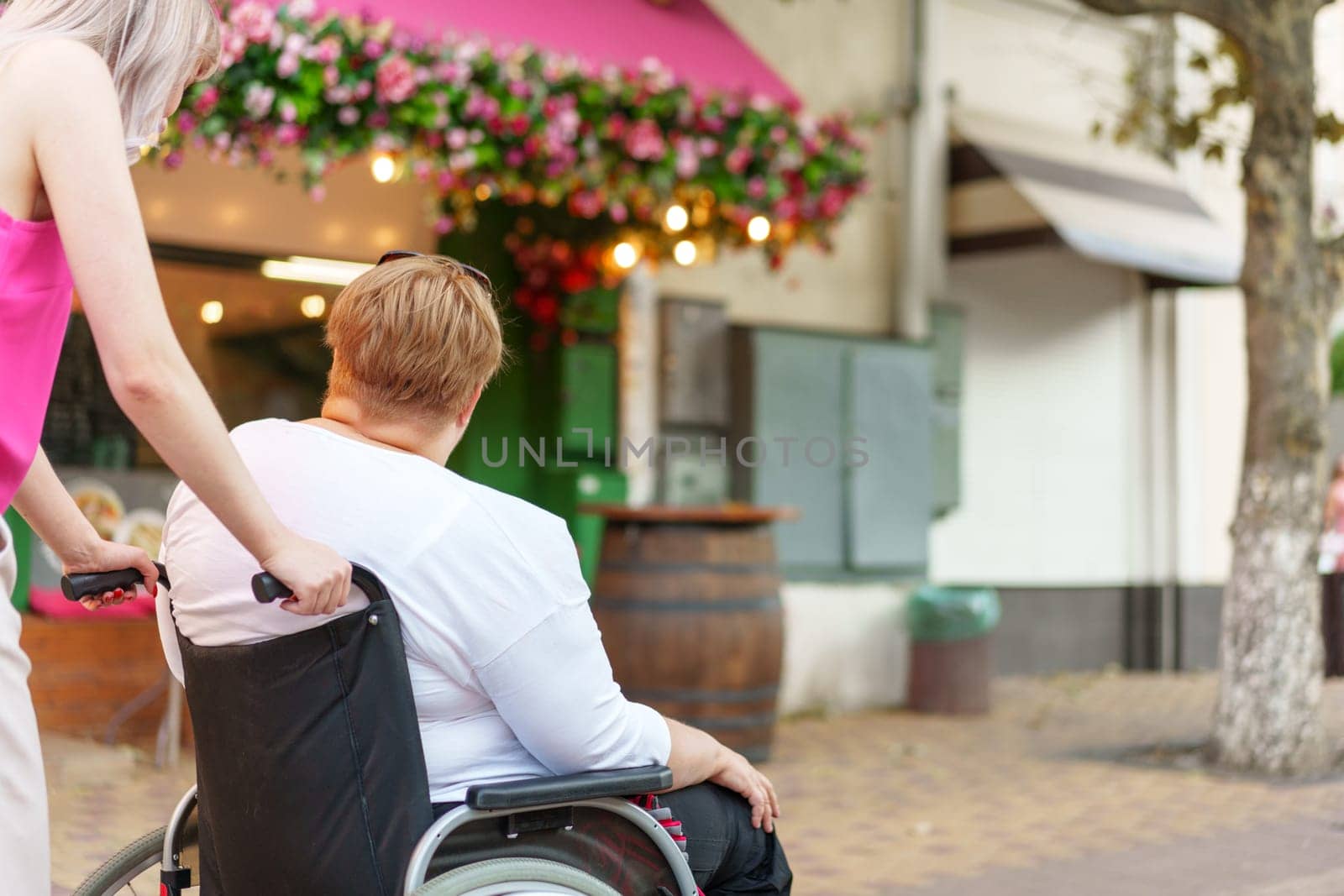 Back view of young woman helping mature woman in wheelchair strolling in the city