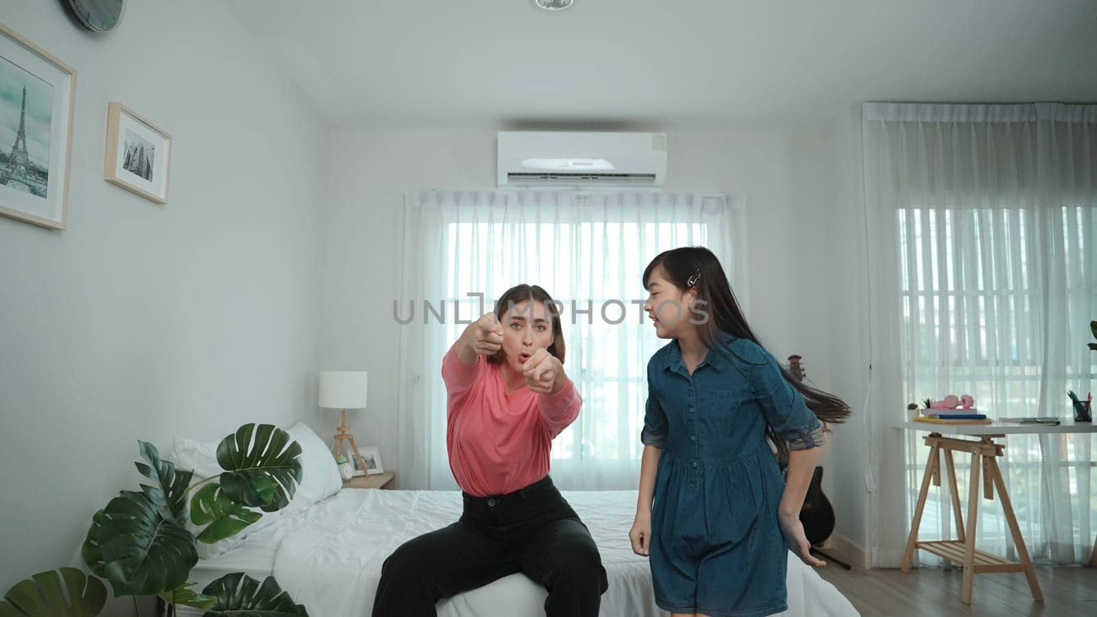 Happy attractive mother dancing to music together with asian daughter at bedroom. Caucasian mom dance or exercise while cute child move to music while jump to bed. Family recreation concept. Pedagogy.