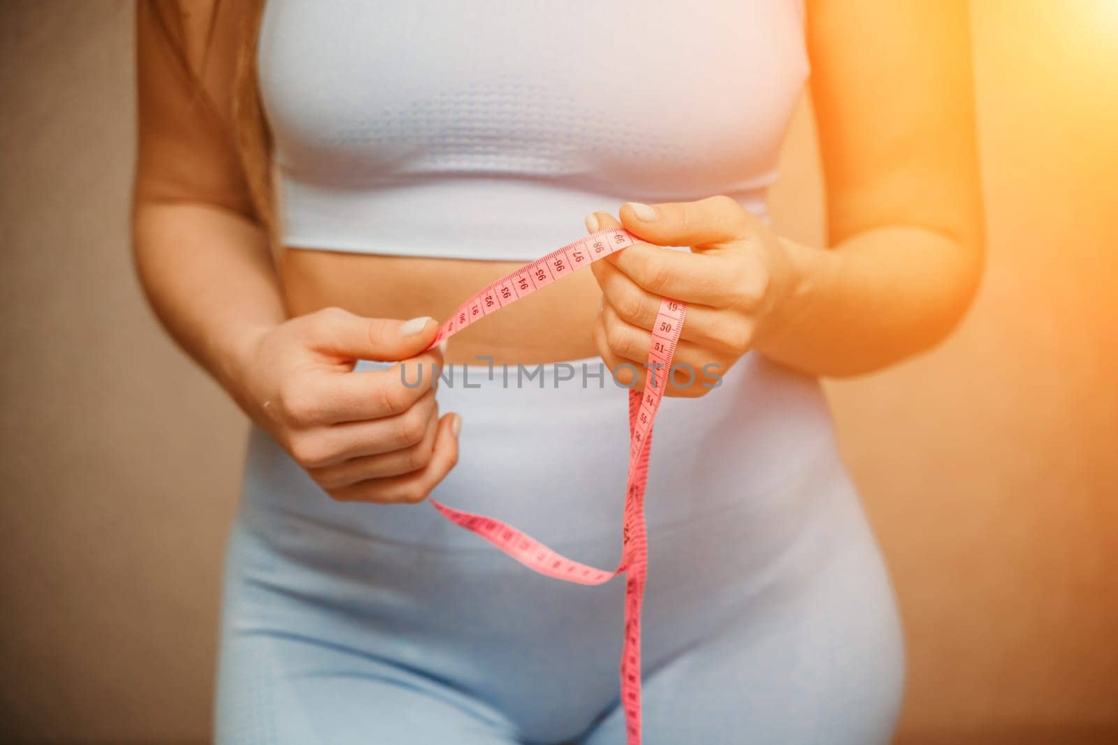 Cropped view of slim woman measuring waist with tape measure at home, close up. European woman checking the result of diet for weight loss or liposuction indoors.