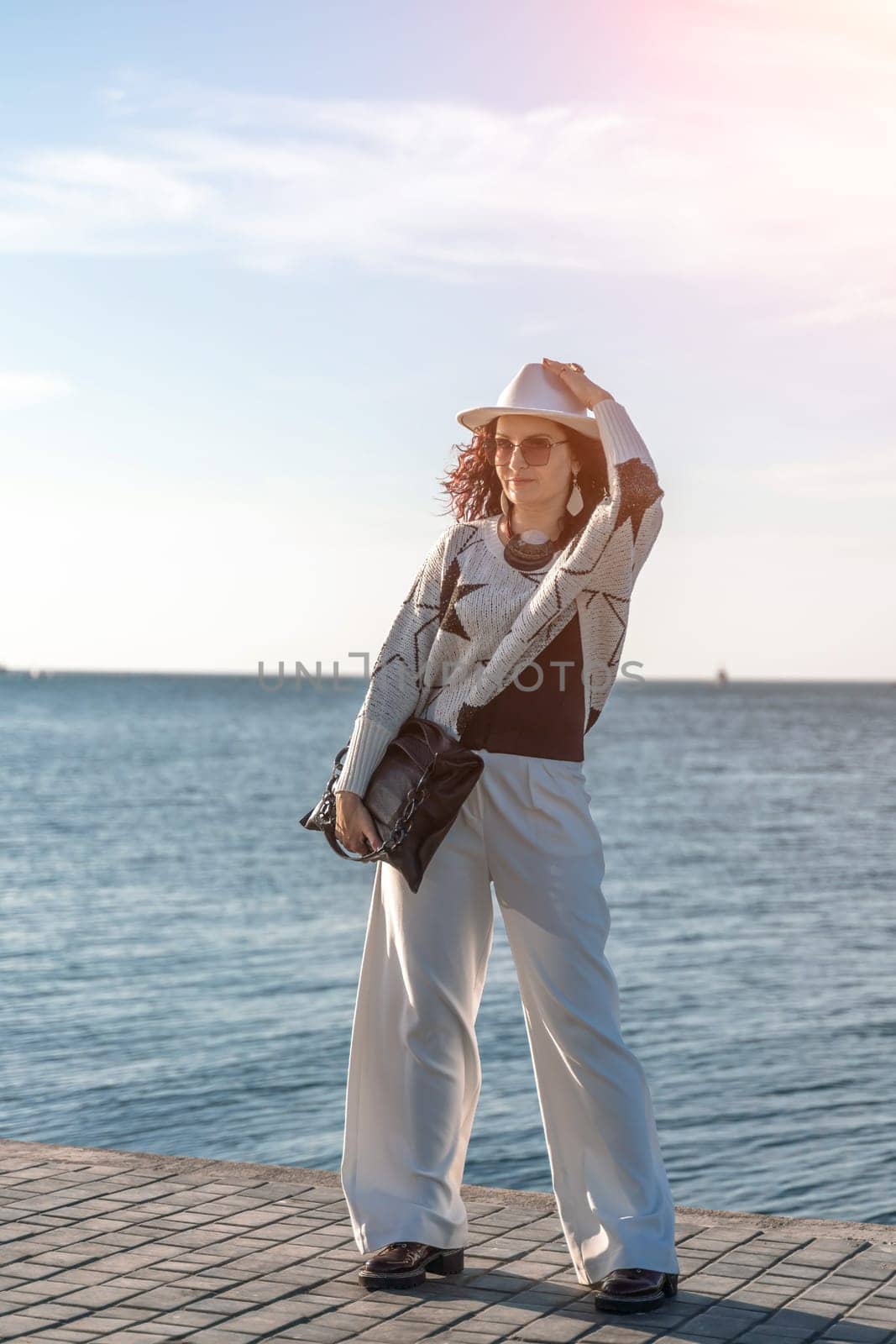 Stylish seashore woman. Fashionable woman in a white hat, white trousers and a light sweater with a black pattern on the background of the sea