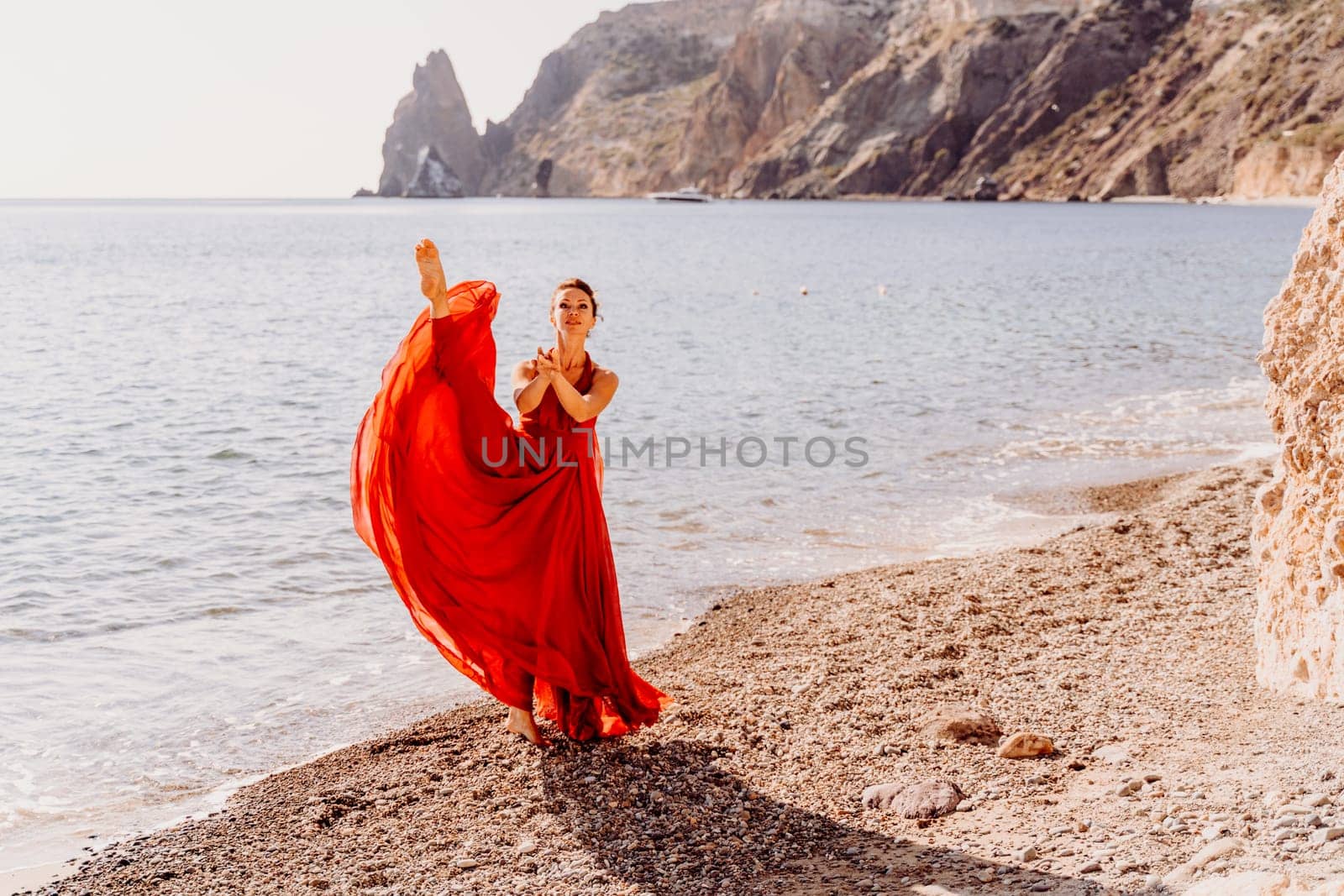 Woman red dress sea. Female dancer in a long red dress posing on a beach with rocks on sunny day. Girl on the nature on blue sky background