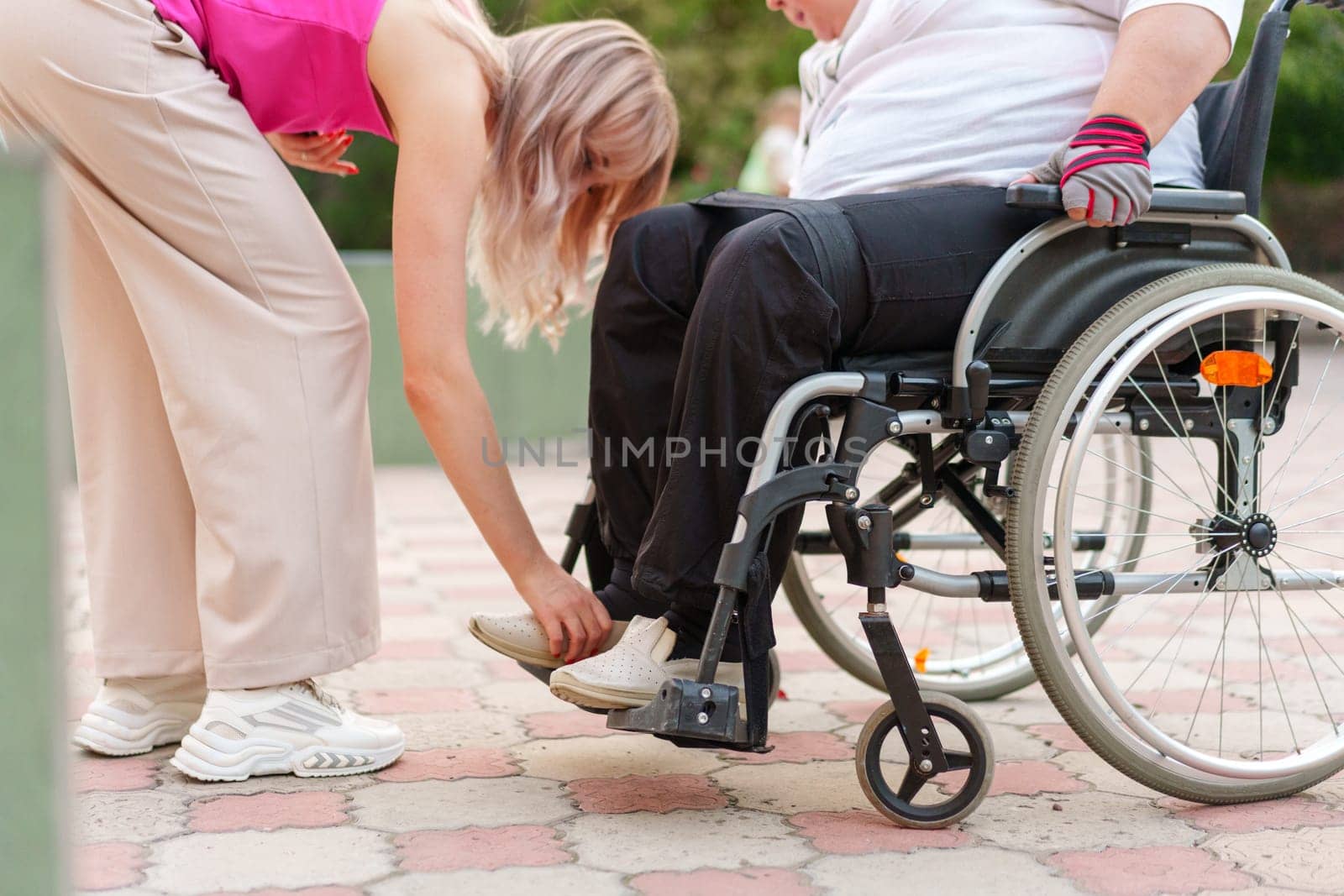 Young unrecognizable female helping woman patient with disability sitting on wheelchair in the street