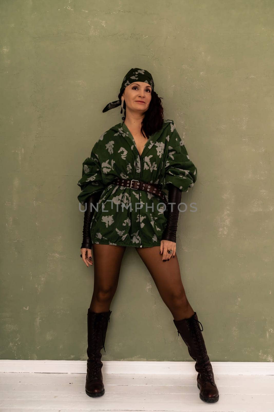 A woman in a green military uniform stands in front of a wall. She is wearing a bandana and has a confident posture