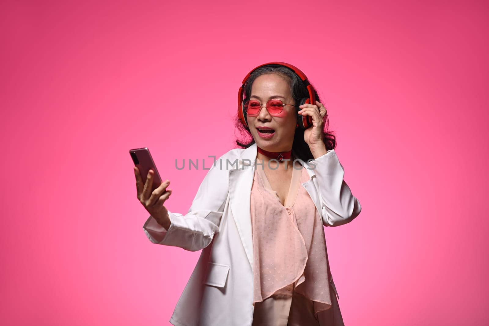 Cheerful retired woman in stylish glasses listening to music in headphones and using mobile phone on pink background.