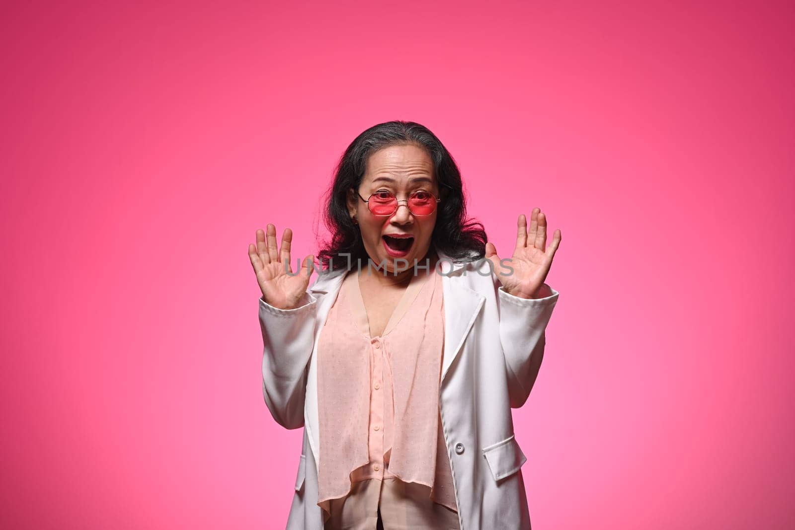 Stylish senior woman standing over pink background with surprise expression.