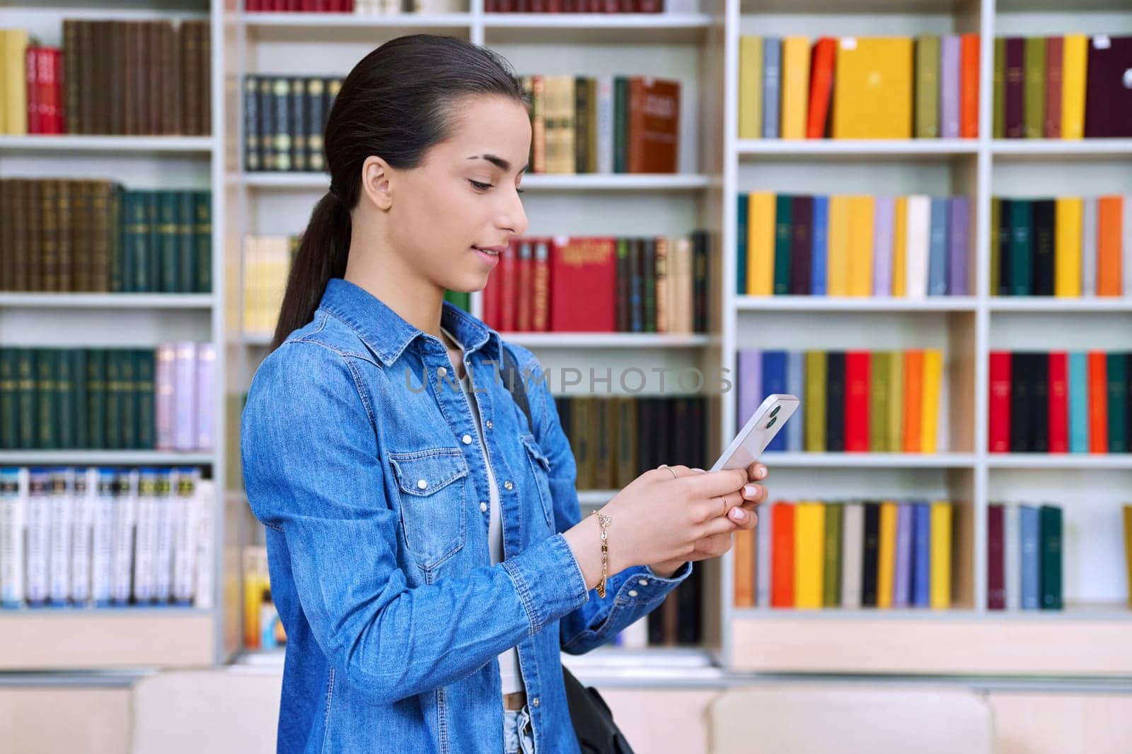 Teenage female student with backpack using smartphone, inside high school building, in library. Technologies, mobile educational apps applications, services, e-learning concept