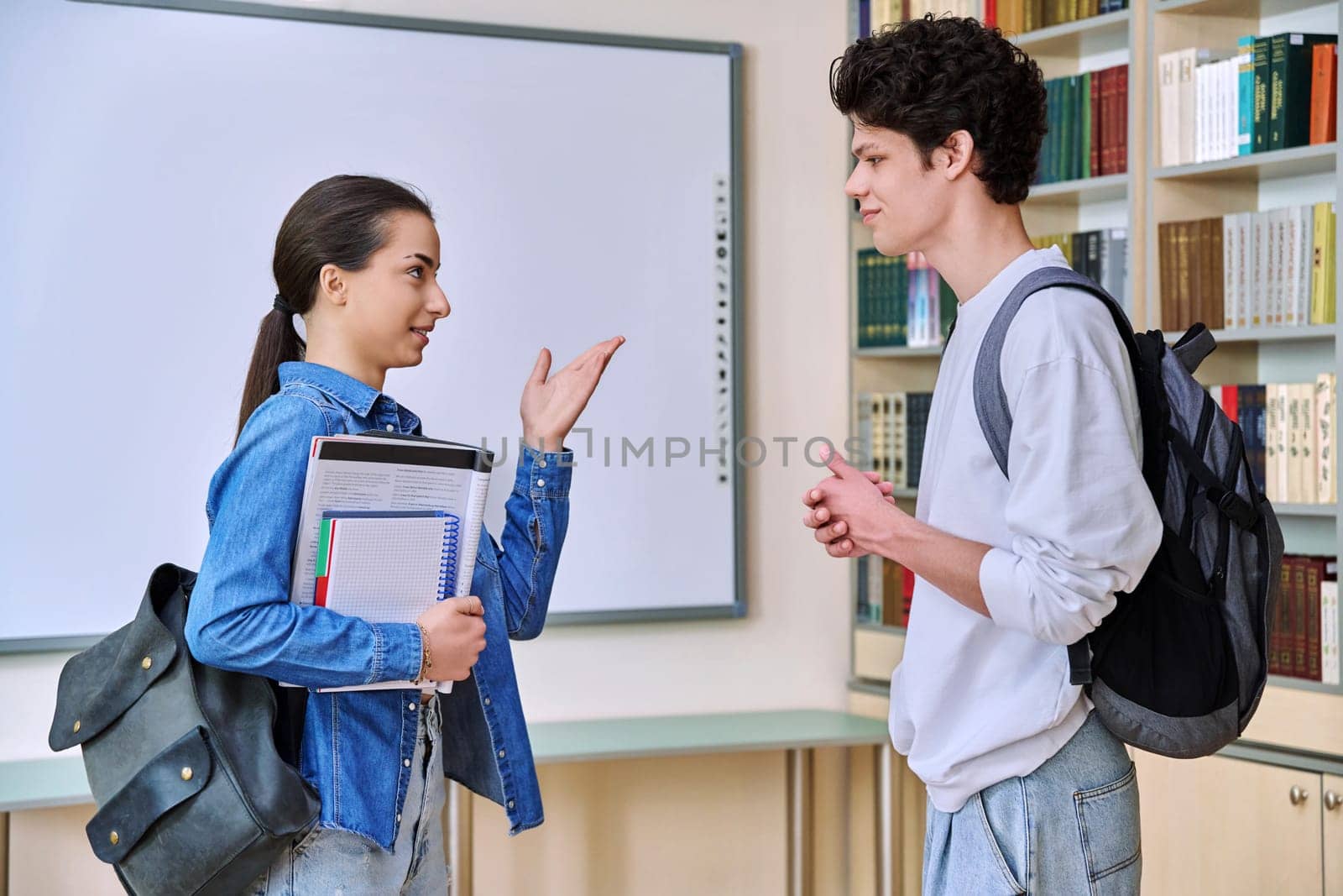 Portrait of two teenage college students classmates, guy and girl, inside library of educational building. Smiling teenage friends talking together. Education, adolescence, youth, lifestyle concept