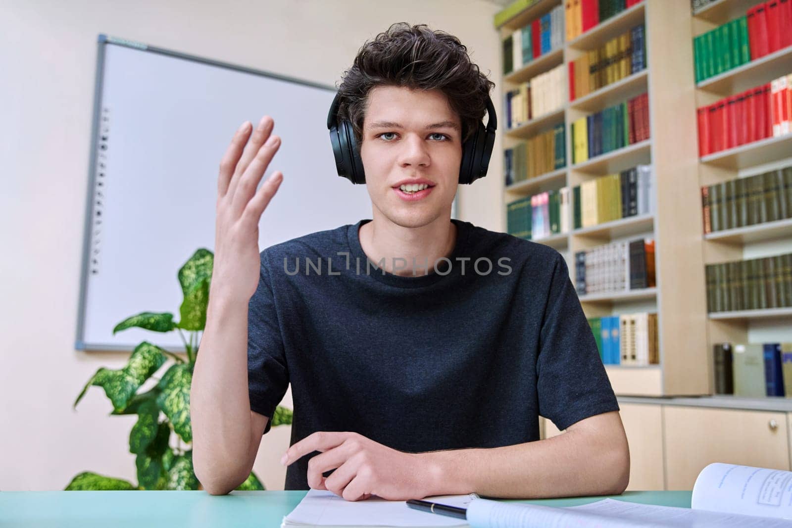 Web cam portrait of college student guy in headphones looking talking to camera in educational building library classroom. Video call chat conference, online lesson exam test, technology education