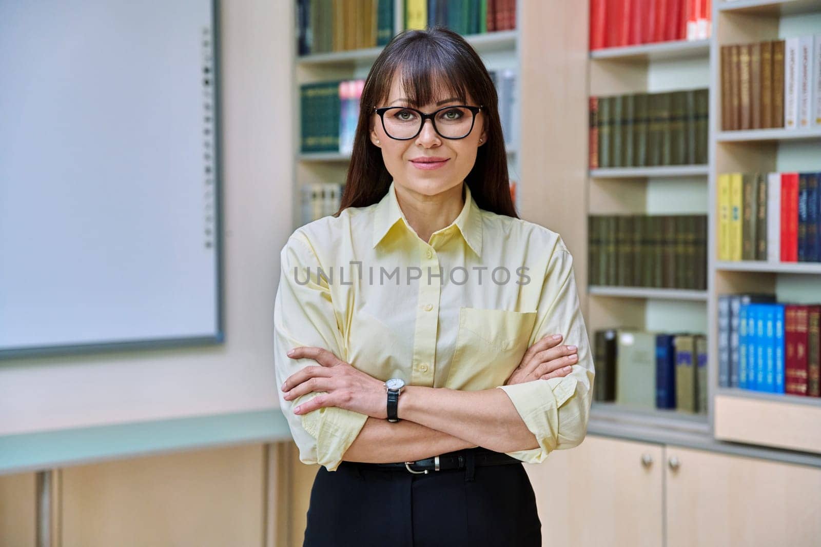 Portrait of confident mature business woman in library. Middle-aged elegant female with crossed arms, manager teacher mentor psychologist counselor advisor librarian background of shelves with books