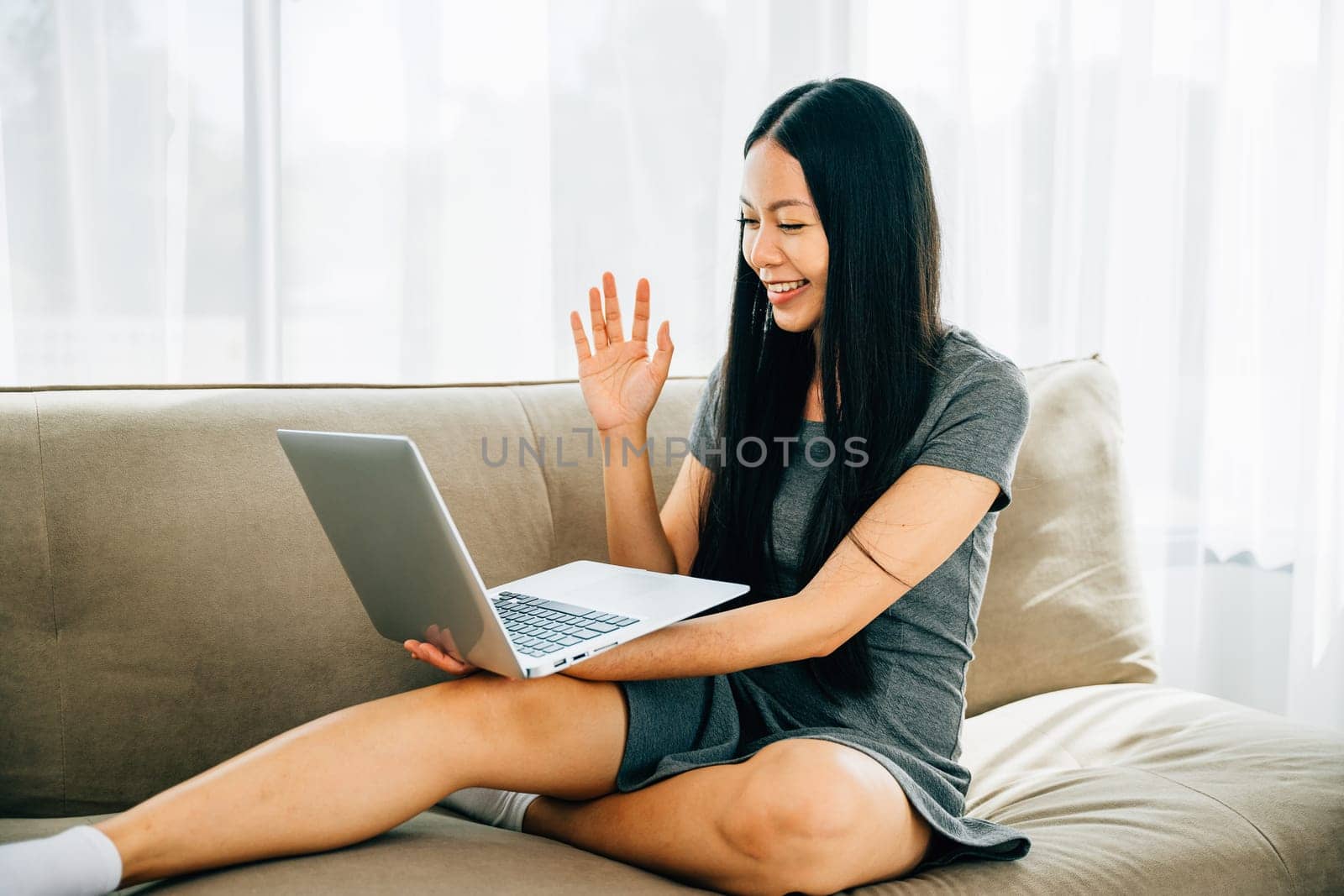 Attractive woman on sofa with laptop waves having video call. Smiling teacher greets welcoming clients. Engaged in distant meeting fostering connections and communication.