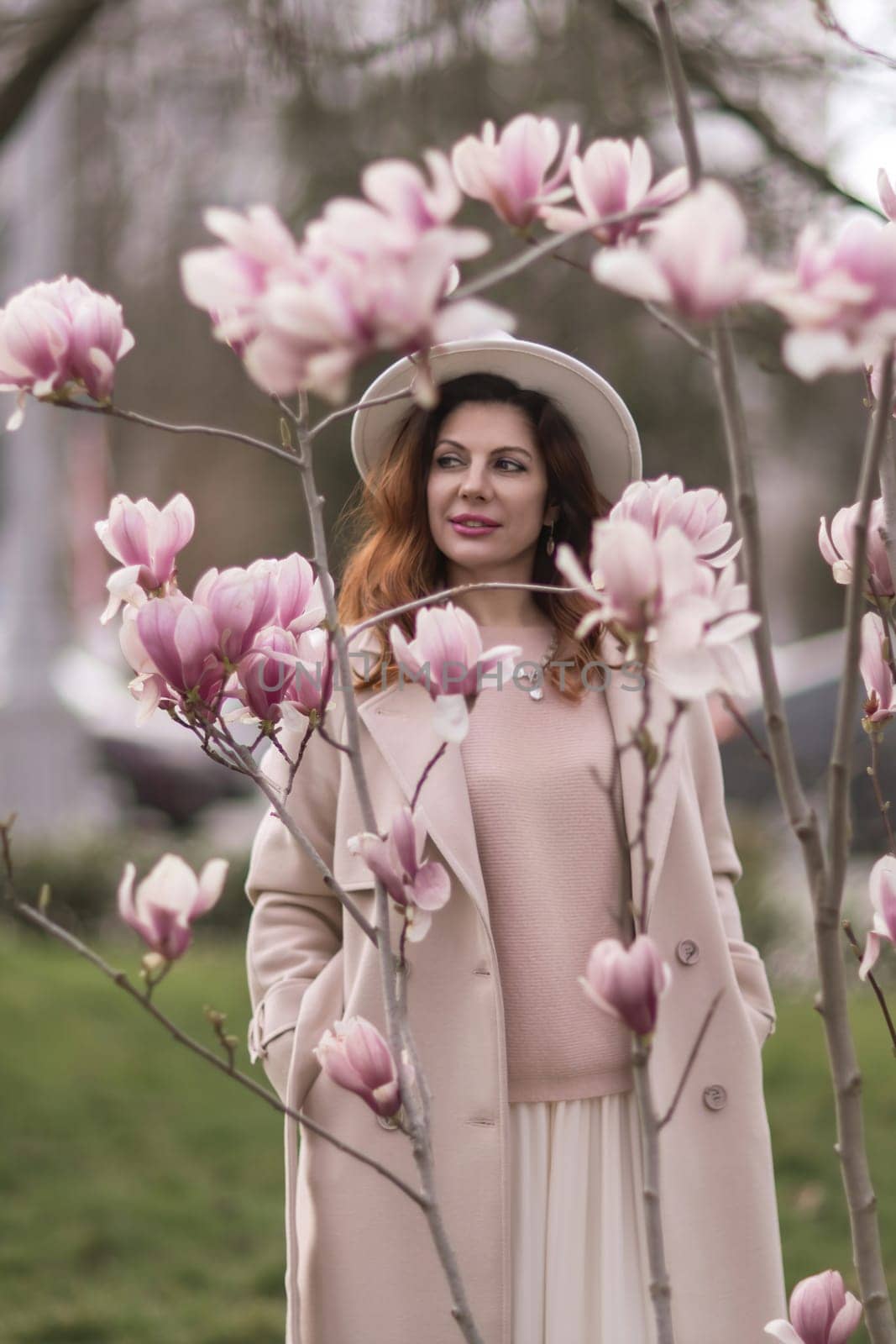 Woman magnolia flowers, surrounded by blossoming trees, hair down, white hat, wearing a light coat. Captured during spring, showcasing natural beauty and seasonal change