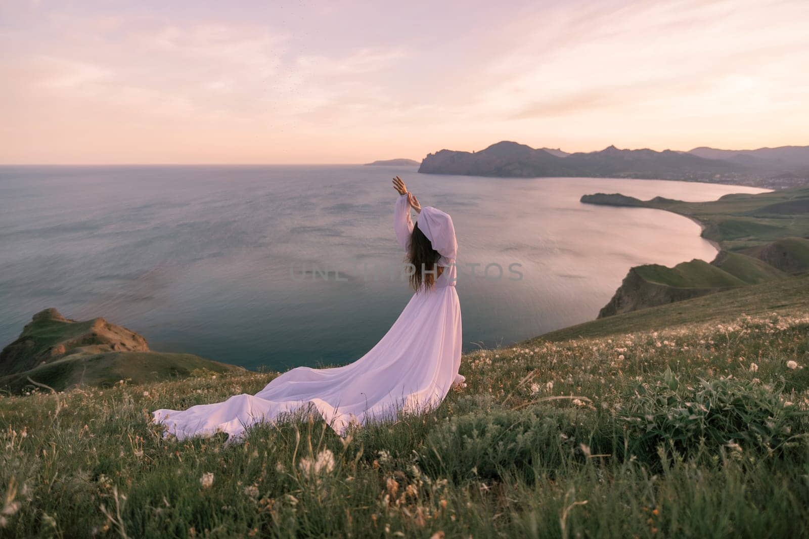 A woman in a white dress stands on a grassy hill overlooking the ocean. The scene is serene and peaceful, with the woman's pose and the beautiful landscape creating a sense of calm and tranquility