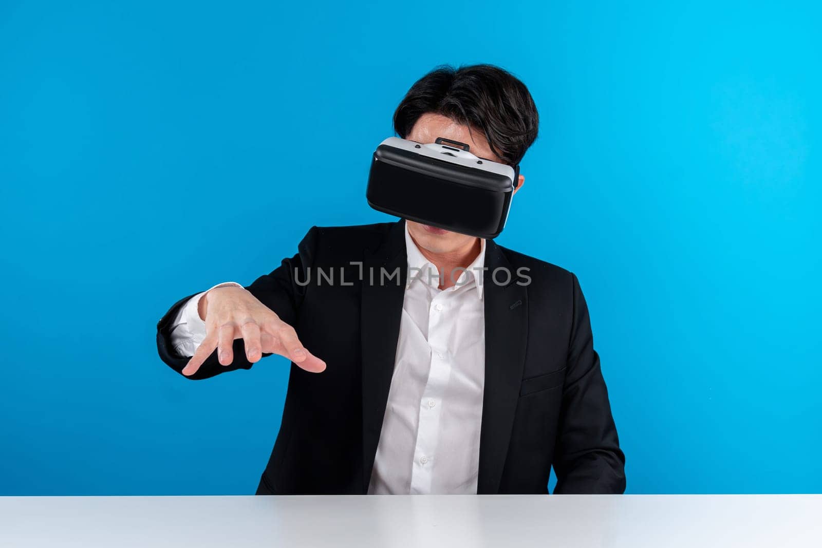 Asian businessman looking through VR connecting meta holding hologram object on table analyzing futuristic business metaverse innovation technology virtual reality isolate blue copyspace. Contrivance.