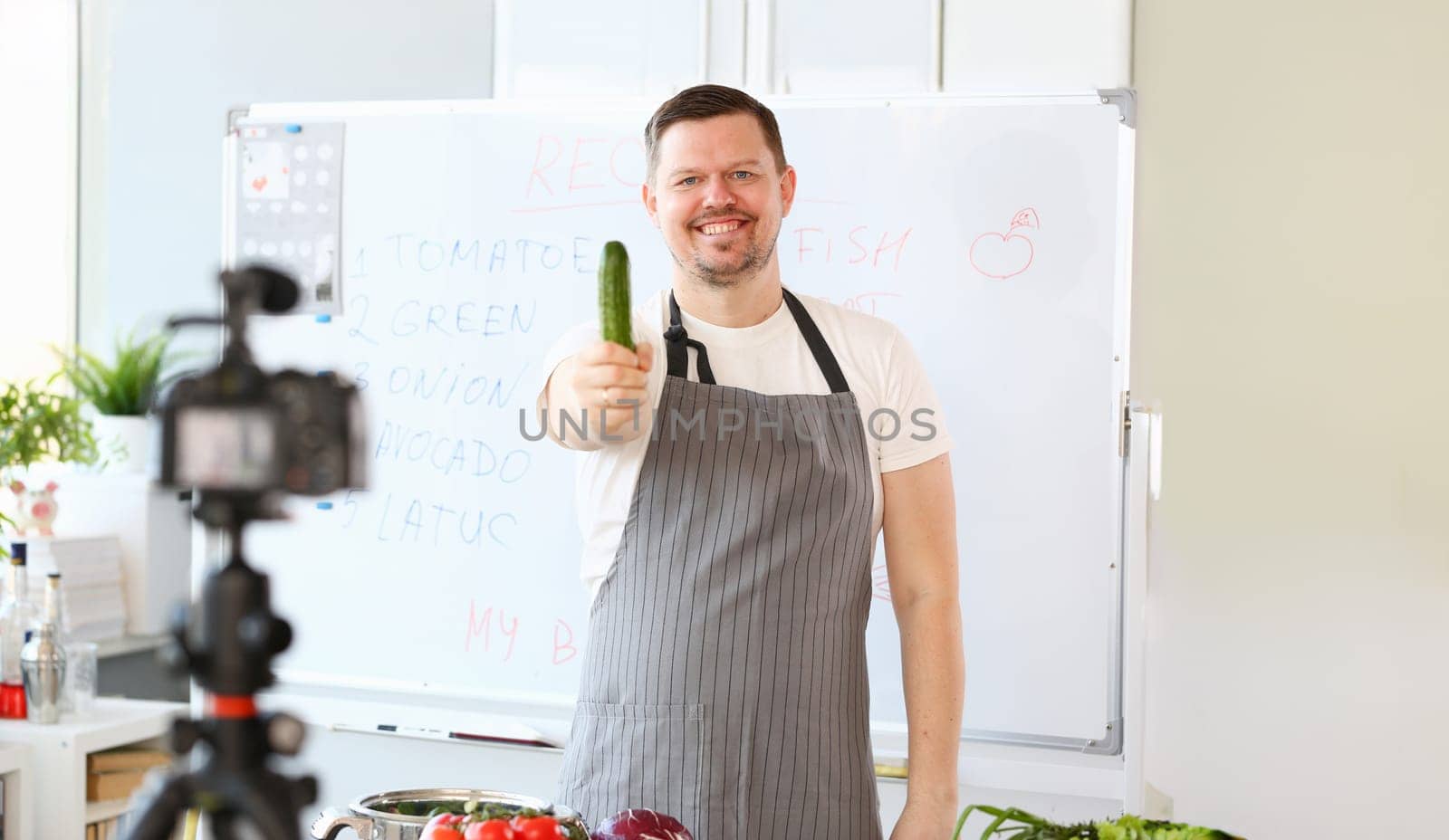 Smiling Vlogger Chef Recording Green Cucumber. Cook in Apron Cooking Vegetable on Camera. Culinary Recipe Vlog at Kitchen. Organic Raw Vegetarian Food. Man with Ripe Ingredient Looking at Camera Shot