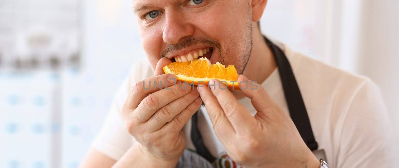 Man Chef Eating Juice Orange Kitchen Portrait. Blogger Person Bite Slice of Citrus Fruit on White Background. Healthy Lifestyle Concept. Cook Holding Fresh Food in Hands Looking at Camera Shot
