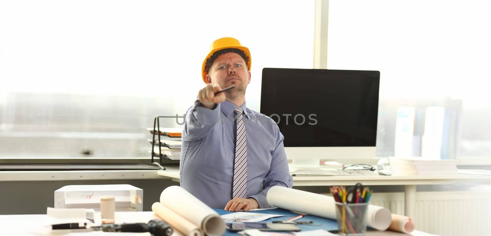 Serious Architect Working on Construction Outline. Caucasian Engineer Sitting at Office Workplace. Man Wear Formal Shirt, Tie and Yellow Helmet do House Drafting. Businessman Pointing at Camera