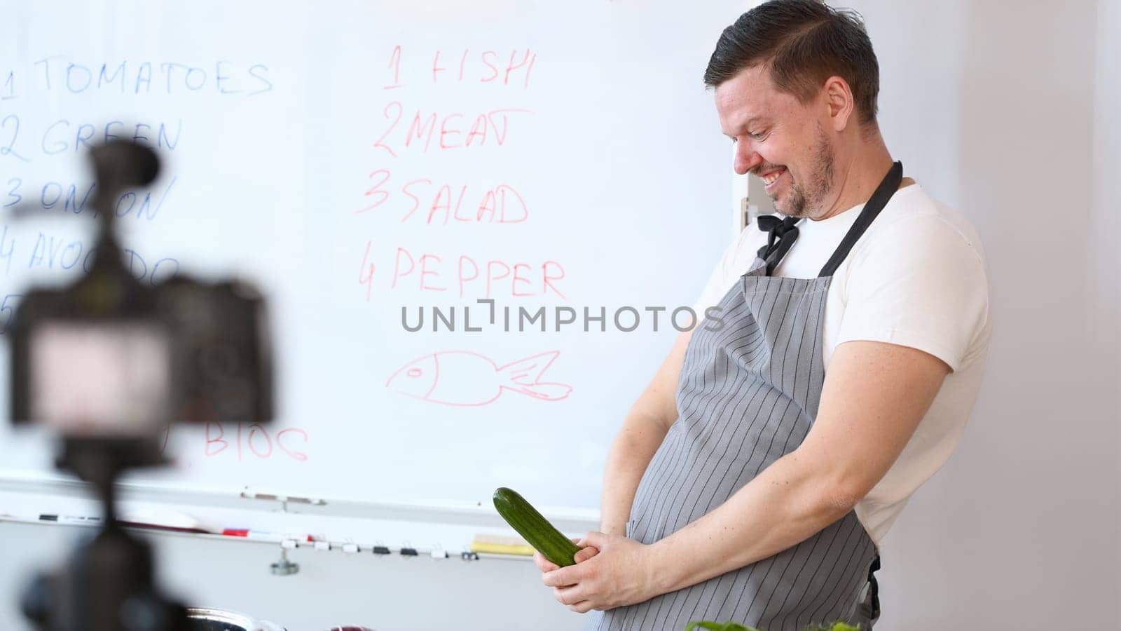 Smiling Vlogger Chef Recording Cucumber Joke. Cook in Apron Joking with Green Cuke on Camera. Culinary Vlog for Vegan at Home Kitchen. Cheerful Man Vegetable Ingredient for Cooking Vegetarian Food