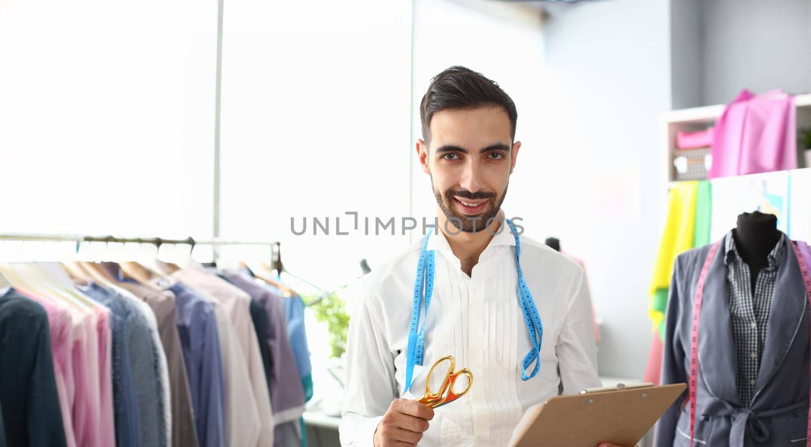 Clothes Creating Work Fashion Atelier Interior. Male Designer Standing with Scissors, Holding Clipboard. Handsome Bearded Man Dressmaker with Measuring Tape over Neck. Mannequin on Background