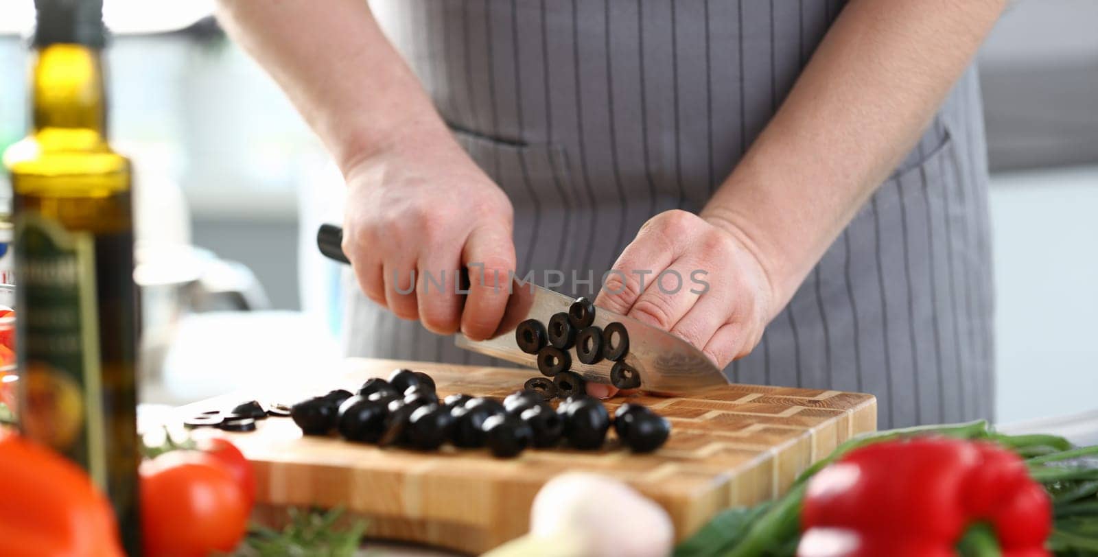 Professional Chef Cutting Black Olive Ingredient. Man in Apron Chopping by Big Kitchen Knife on Wooden Board. Healthy Culinary Recipe. Cooking Dieting Food at Kitchen Horizontal Photography