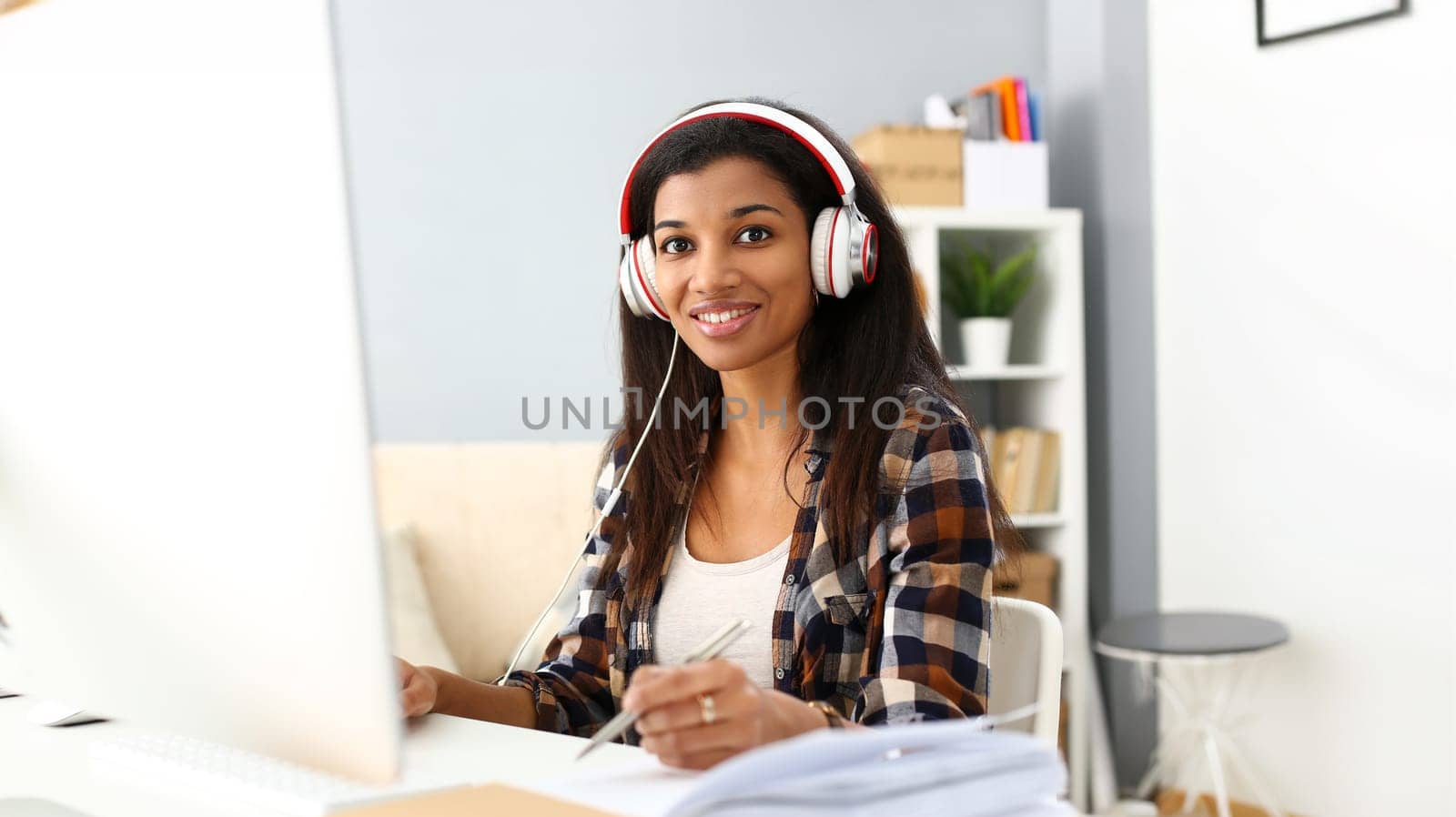 Black smiling woman sitting at workplace wearing headphones studying or doing school homework concept