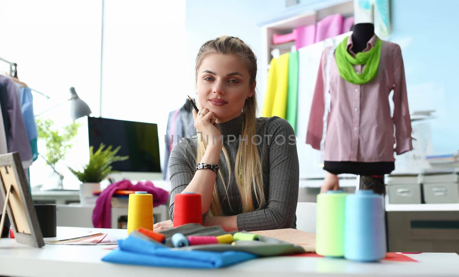 Fashion Clothes Design Handicraft Sewing Concept. Woman Dressmaker at Manufacturing Workplace. Caucasian Beautiful Girl Holding Pen. Fabric, Sewing Tools and Reels of Color Thread on Table