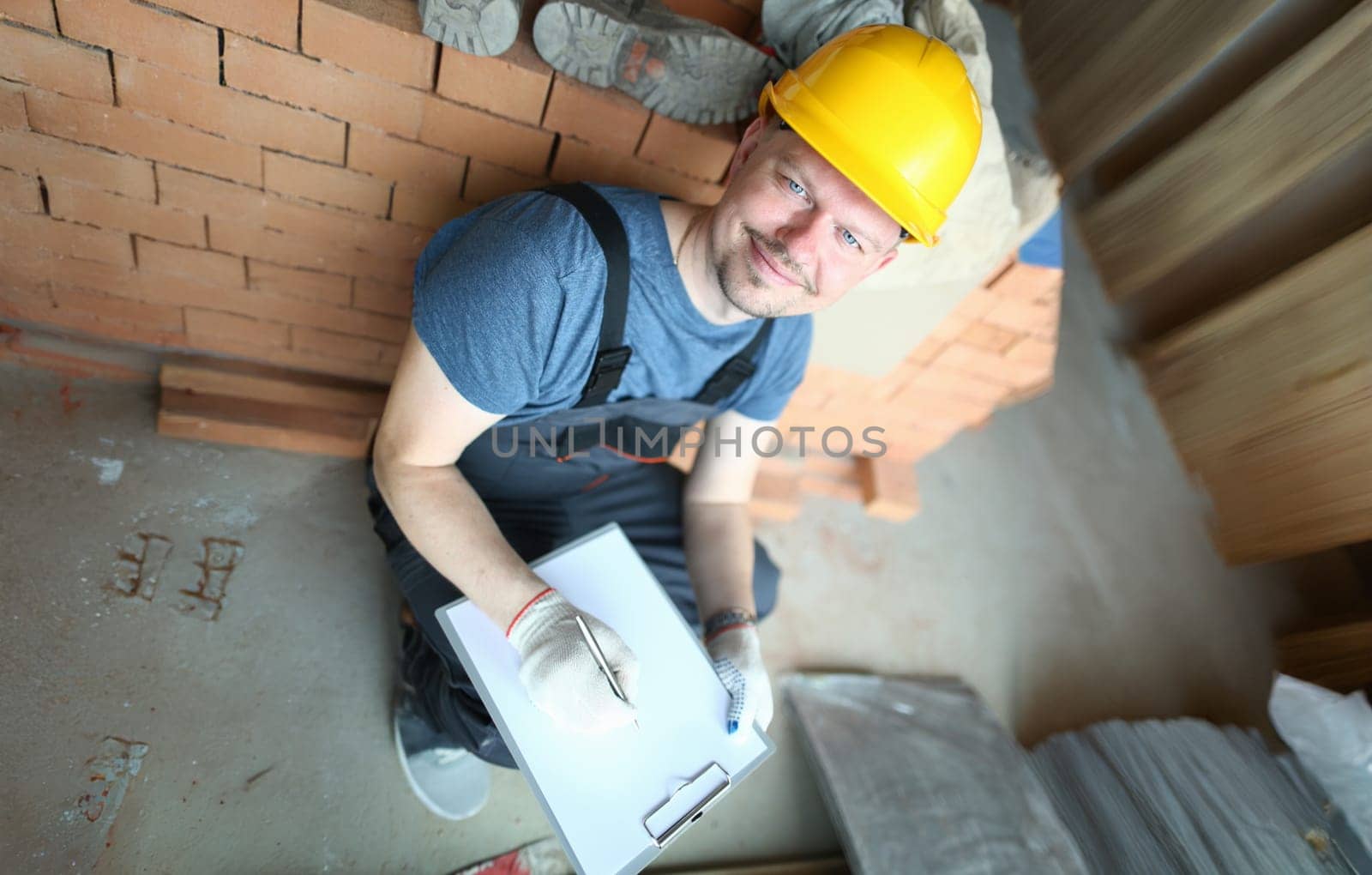 Male builder in yellow hard hat holds clipboard on background of an apartment under repair portrait. Estimated property value concept.