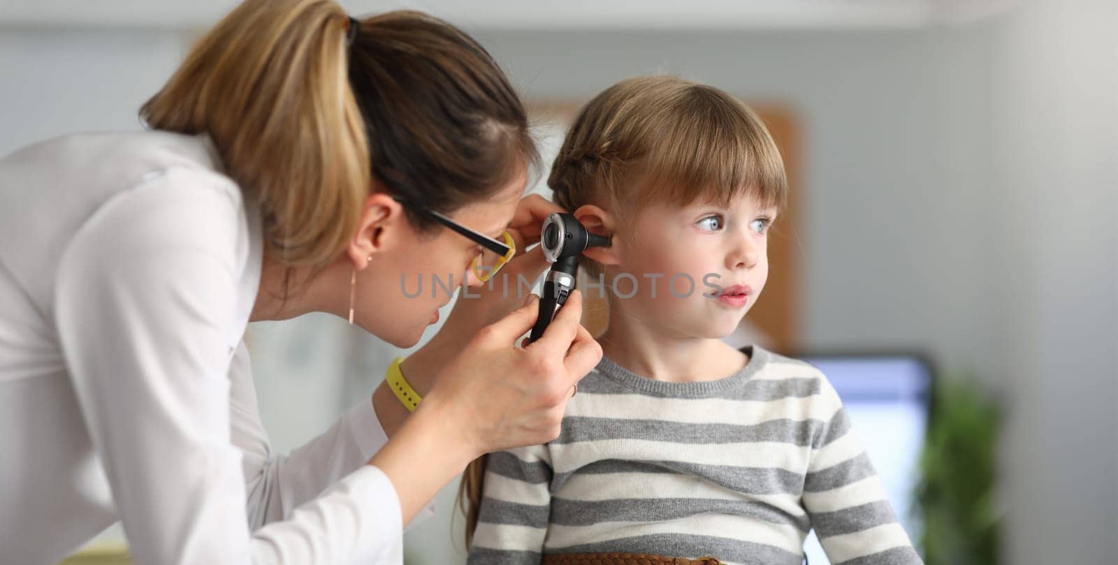 Portrait of smiling girl sitting in modern hospital office of professional physician looking into ear with special medical utility. Health checkup concept