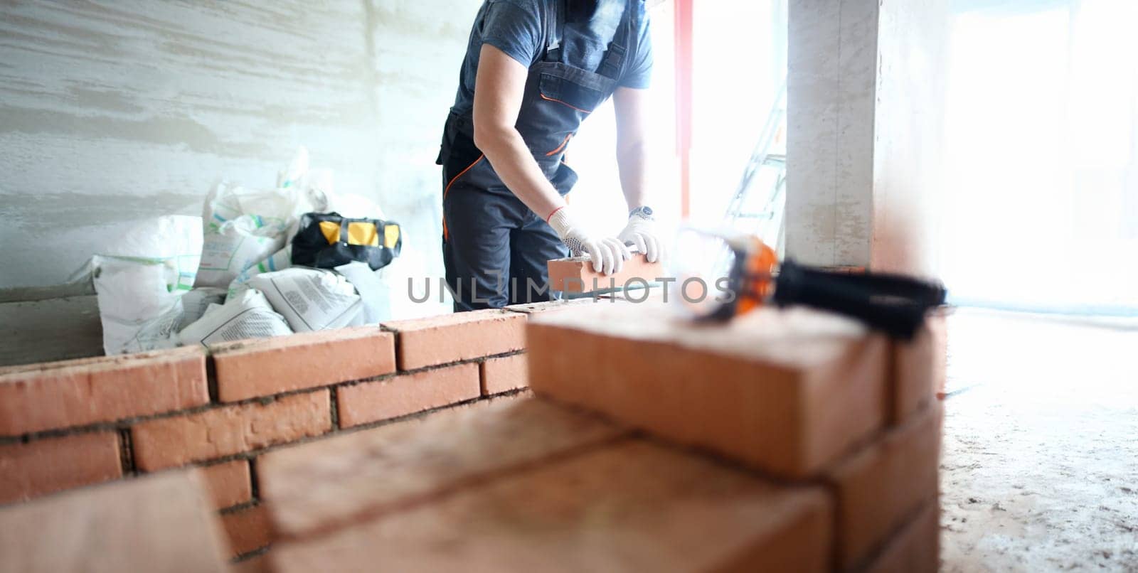 Male builder hand in gloves holding clay brick on background of brickwork wall. Repair and redevelopment apartments concept.