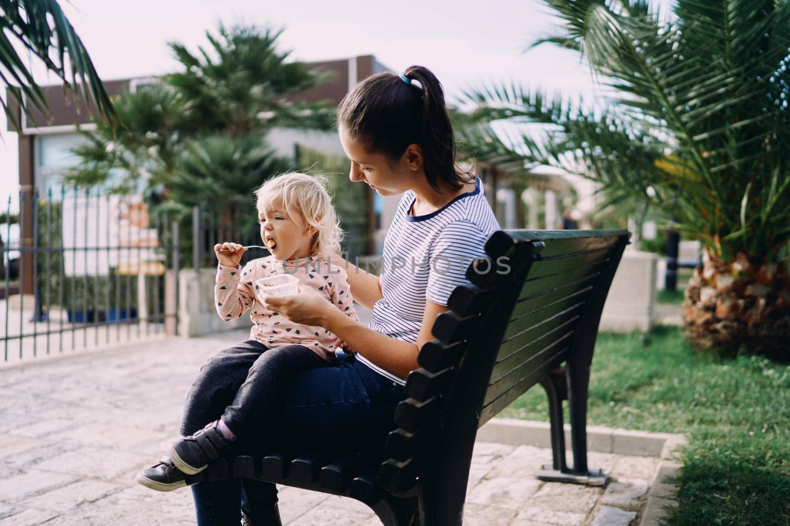 Little girl with a spoon eats porridge from a lunchbox in the hands of her mom sitting on a bench. High quality photo