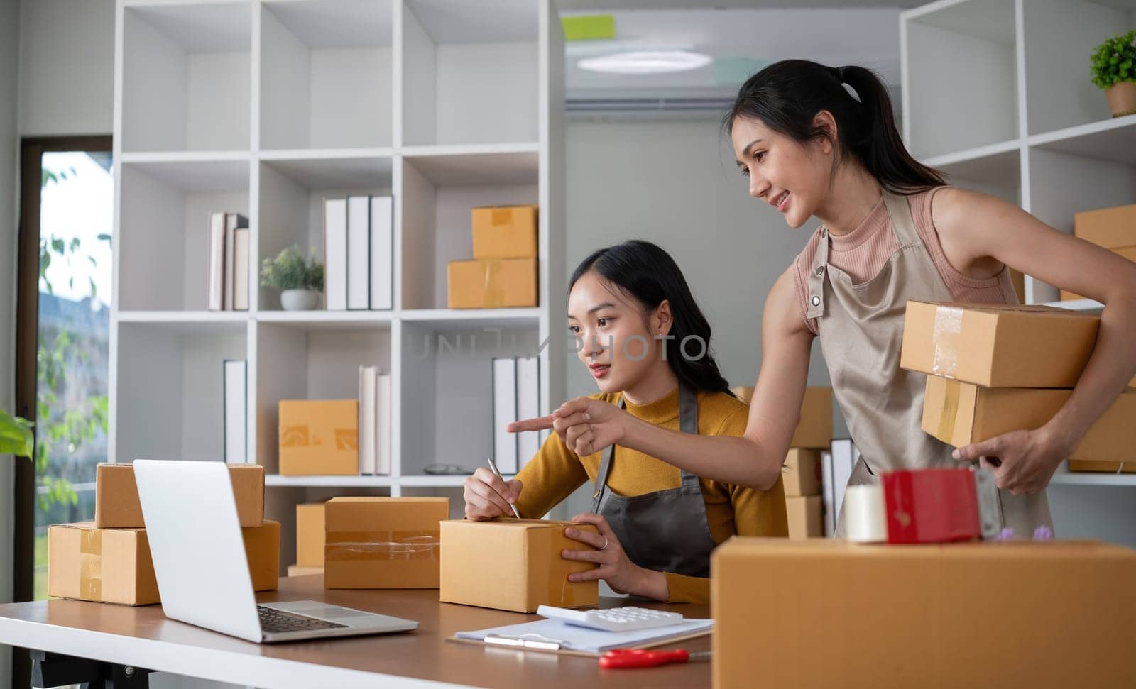 Couple of young Asian women running a small business together selling products online using laptops by wichayada