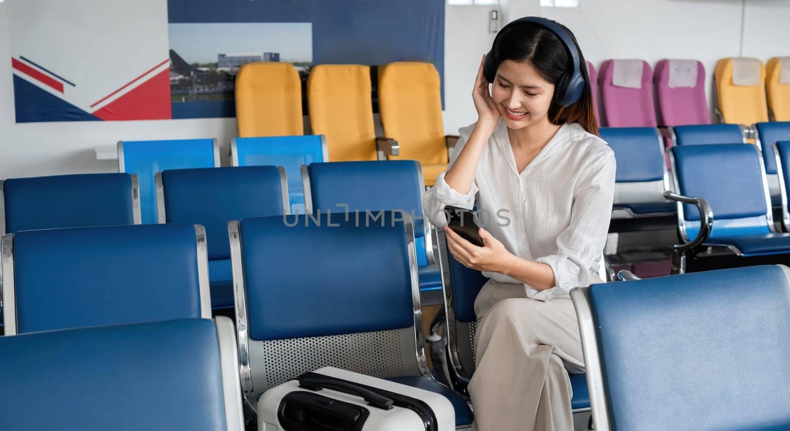 Asian woman listening to music on smartphone in airport waiting area. Concept of travel, technology, and relaxation.