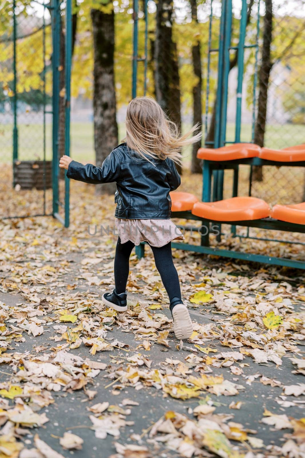 Little girl runs across the sports ground towards the gate, past the rows of seats. Back view. High quality photo