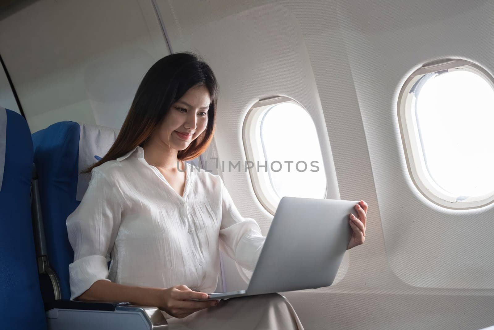 Asian woman working on laptop in airplane seat. Concept of air travel, remote work, and in-flight productivity by wichayada