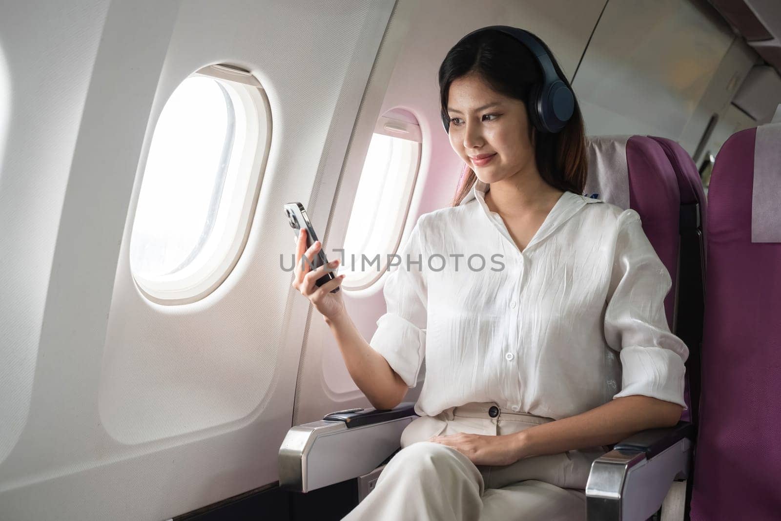Asian woman using smartphone and headphones on airplane. Concept of air travel, technology, and in-flight entertainment.