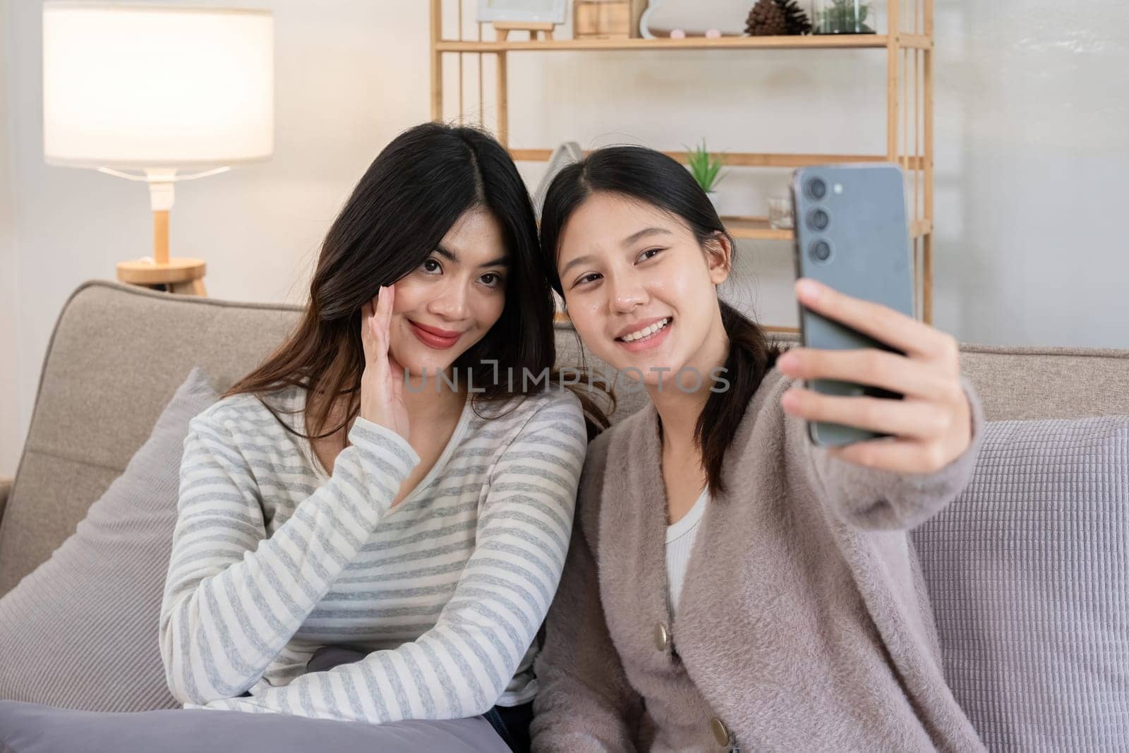 Asian lesbian couple taking a selfie on the sofa at home. Concept of love, togetherness, and capturing happy moments in a cozy indoor setting.