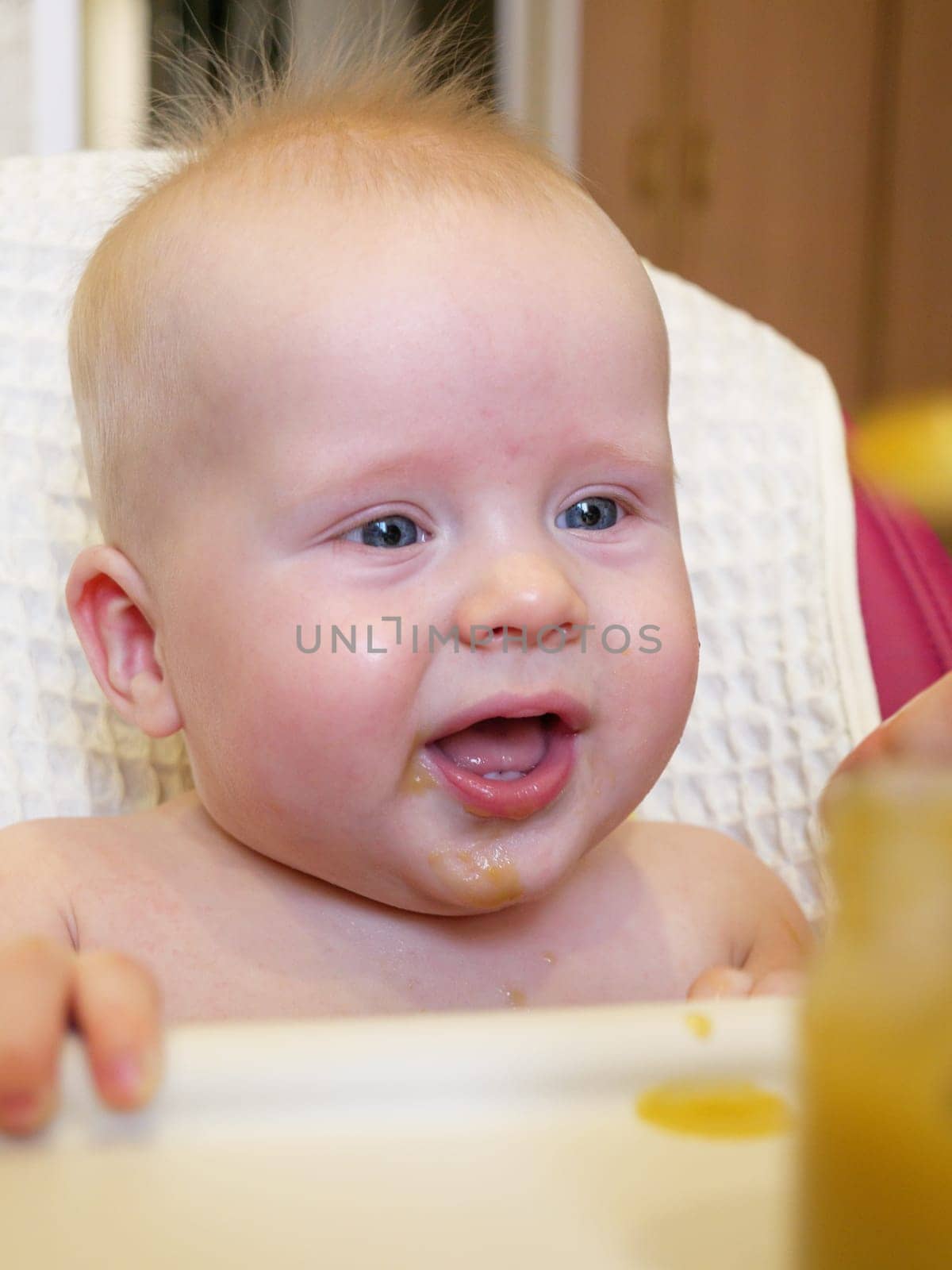 A cute baby with blue eyes has broccoli puree smeared all over their face. It looks like the baby is enjoying their meal while sitting at home.