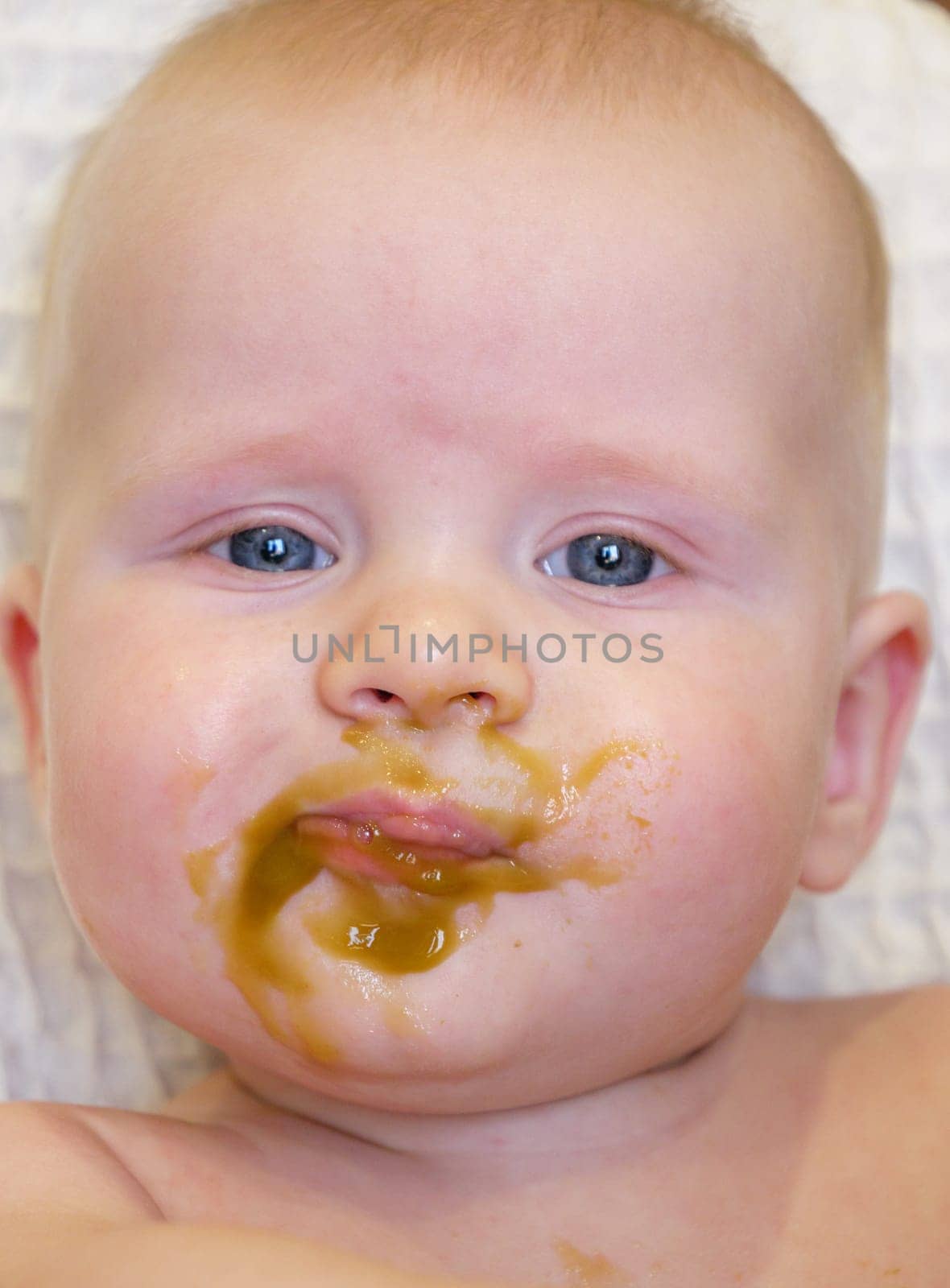 A cute baby with blue eyes has broccoli puree smeared all over their face. It looks like the baby is enjoying their meal while sitting at home.