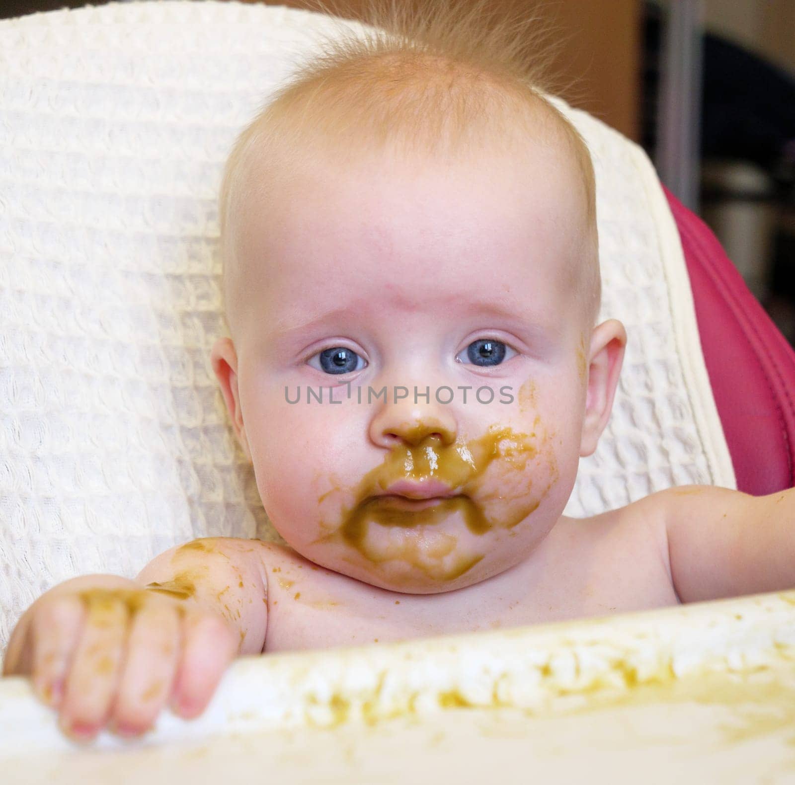 A cute baby with blue eyes has broccoli puree smeared all over their face. It looks like the baby is enjoying their meal while sitting at home.