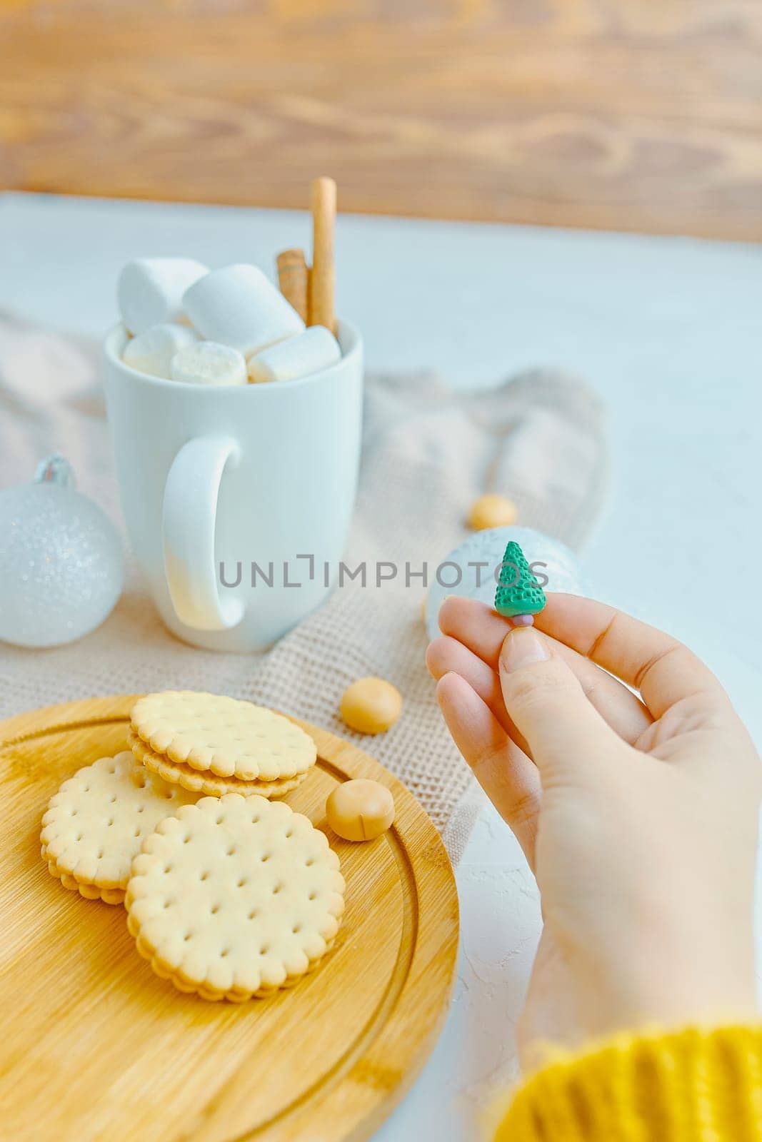 Woman in a yellow sweater drinks hot cocoa with marshmallow and cinnamon rolls in a white mug and cookies and a figure of a Christmas tree