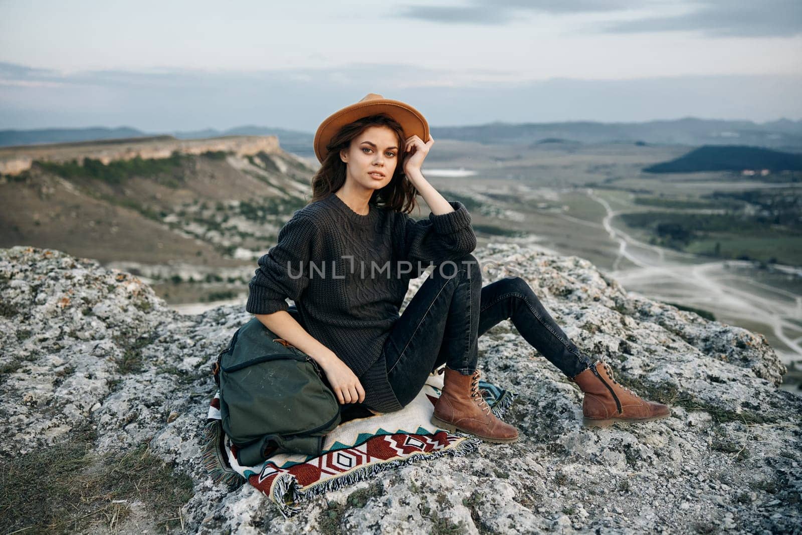 Adventurous woman with backpack and hat sitting on rock at mountain summit
