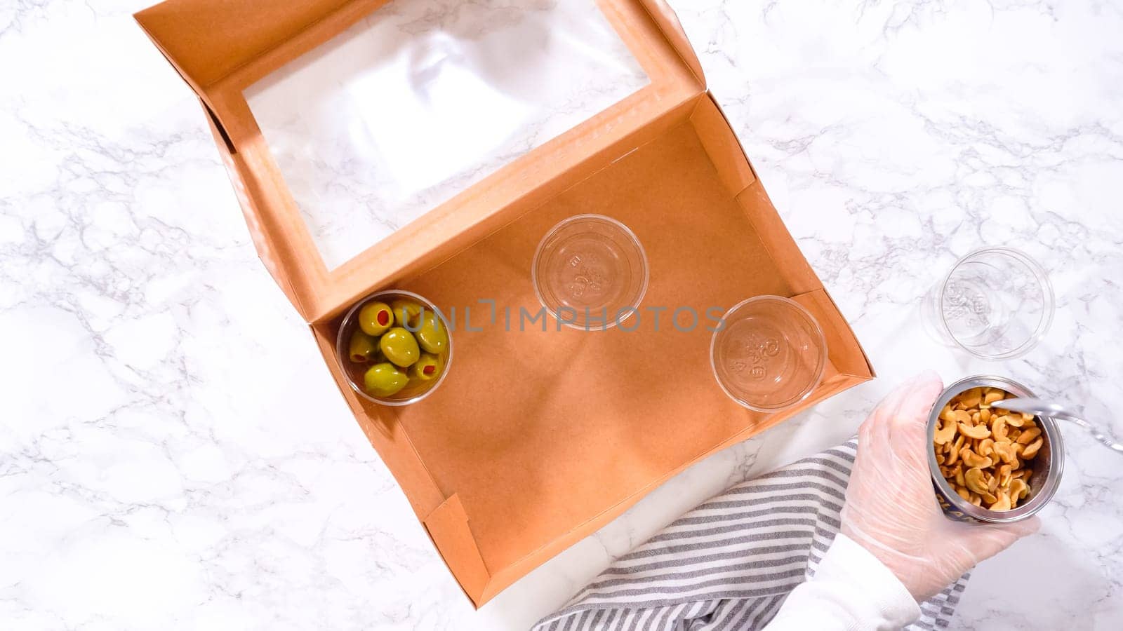 Flat lay. Hands are meticulously adding fresh red grapes to a bowl, complementing a beautifully arranged charcuterie box brimming with a variety of cheeses, olives, and cured meats, set against a sleek marble surface.